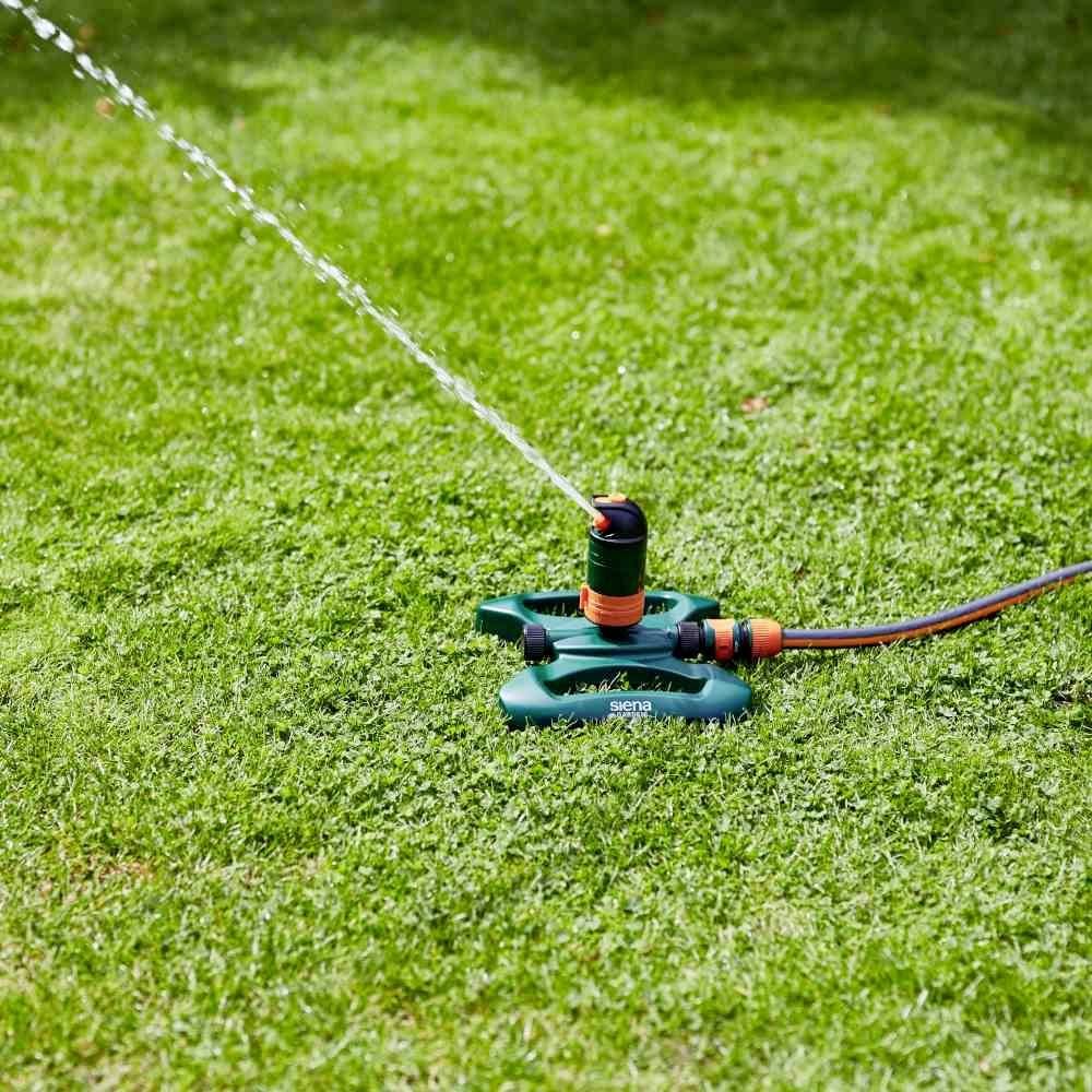 Zraszacz nawadnia zieloną trawę. Strumień wody tryska ukośnie z dyszy natryskowej w powietrze.