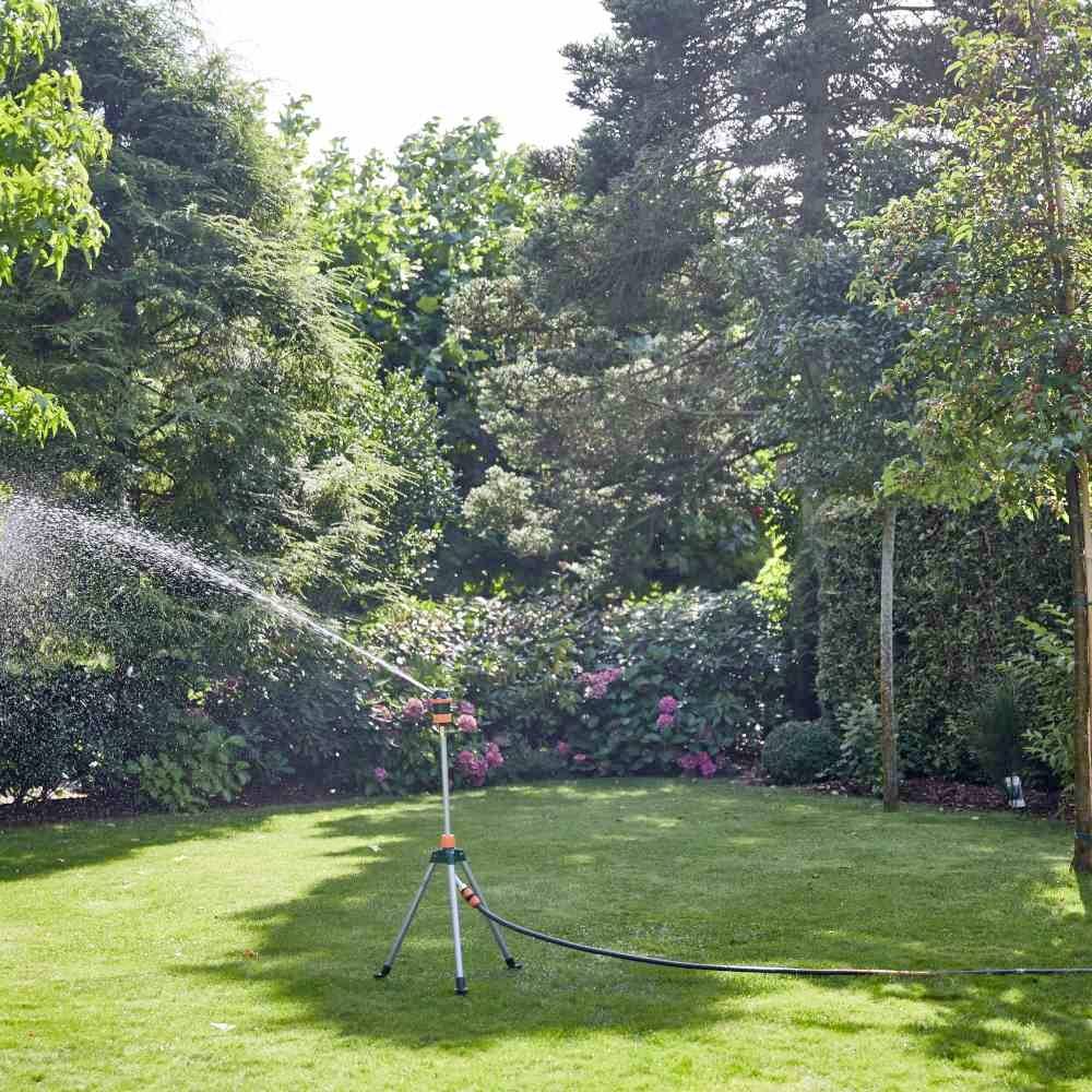 Zraszacz nawadnia zieloną trawę w gęsto porośniętym ogrodzie w świetle dziennym. Wokół sceny rosną różne rośliny ogrodowe.