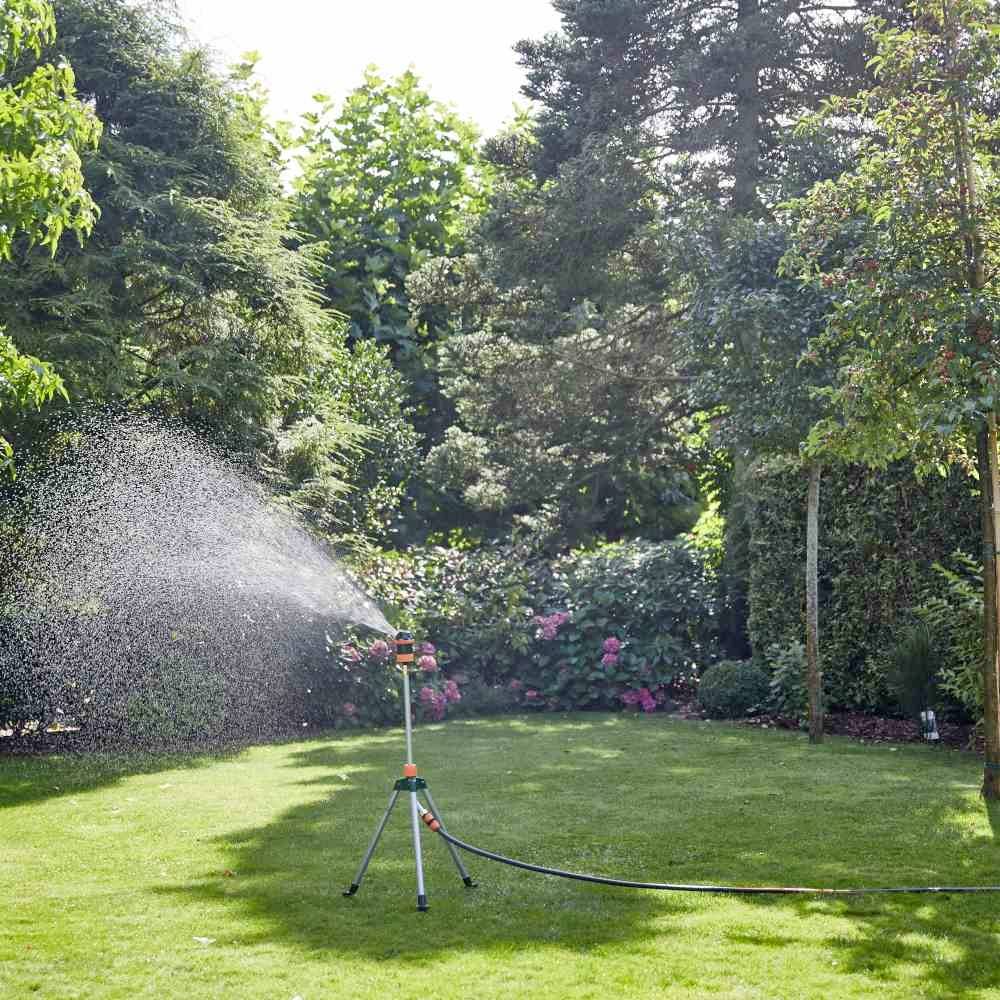 Zraszacz nawadnia zielony trawnik w ogrodzie. Otoczony gęstymi drzewami i krzewami w słoneczny dzień.