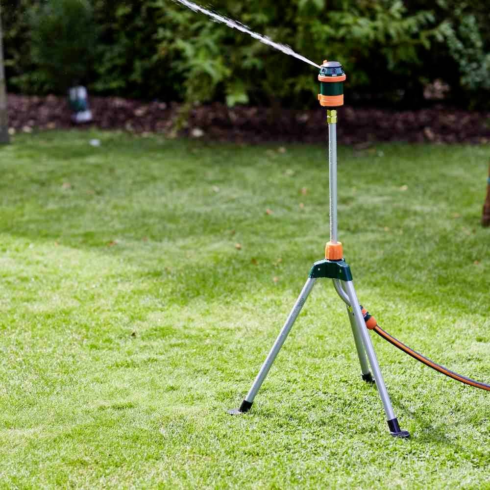 Zraszacz ogrodowy na jednej nodze równomiernie podlewa trawnik wodą. W tle widać drzewa i krzewy.