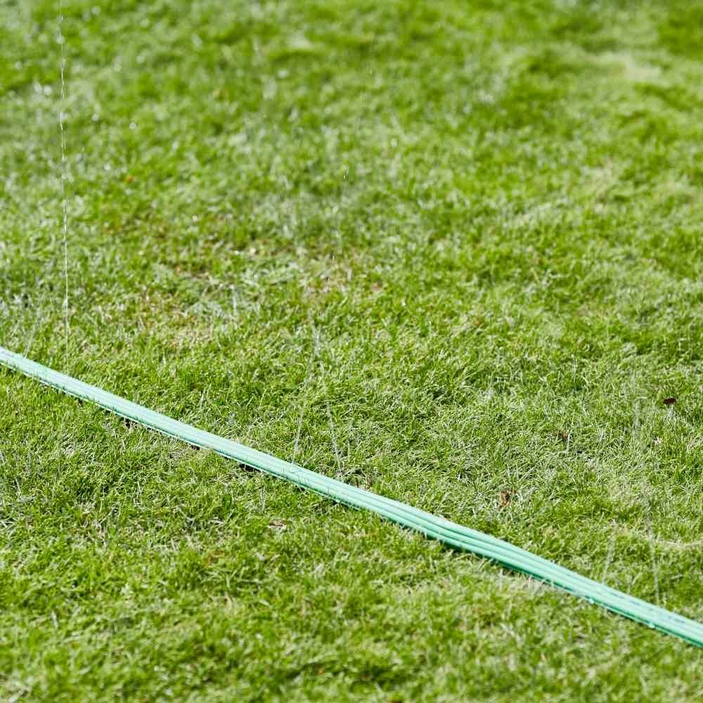 Zielony wąż ogrodowy leży na trawniku i rozpyla drobną mgiełkę wodną, nawilżając trawę.