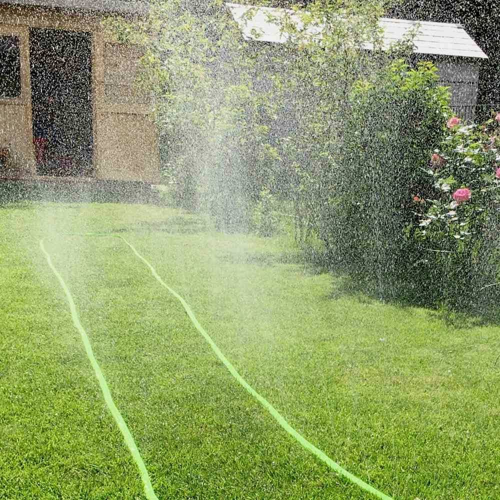 Trawnik z zraszaczem, który rozpyla drobno rozproszony strumień wody. W tle mały schowek i kwitnące krzewy.