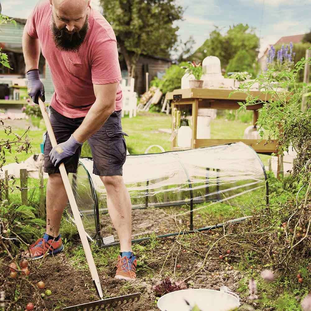 Osoba z brodą pracuje w ogrodzie, ubrana w czerwone ubranie i spulchnia ziemię grabiami. W tle widać rośliny i meble ogrodowe.