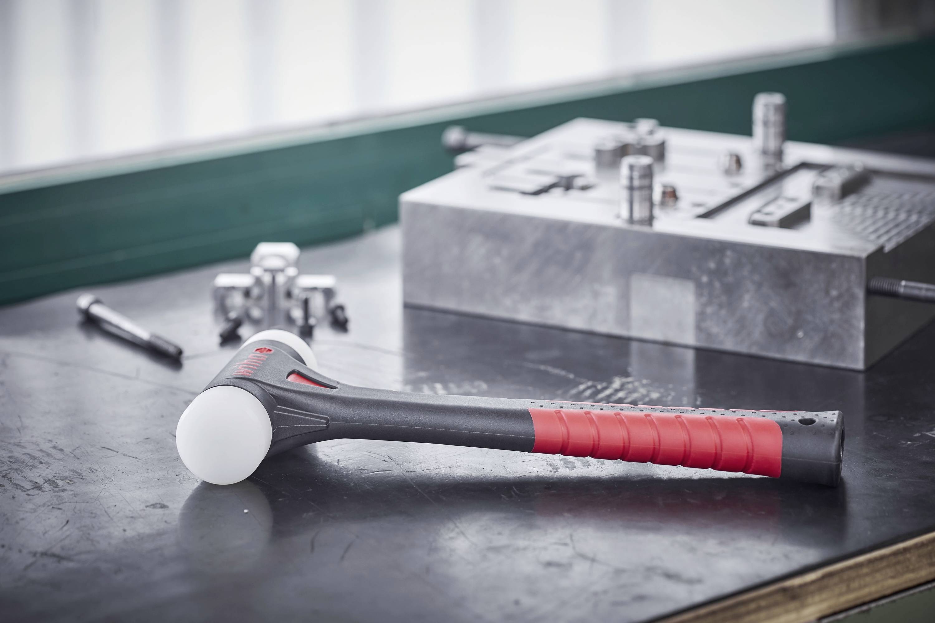 Młotek z czerwoną rączką na stole warsztatowym, otoczony częściami metalowymi i narzędziami, prawdopodobnie w warsztacie lub środowisku produkcyjnym.