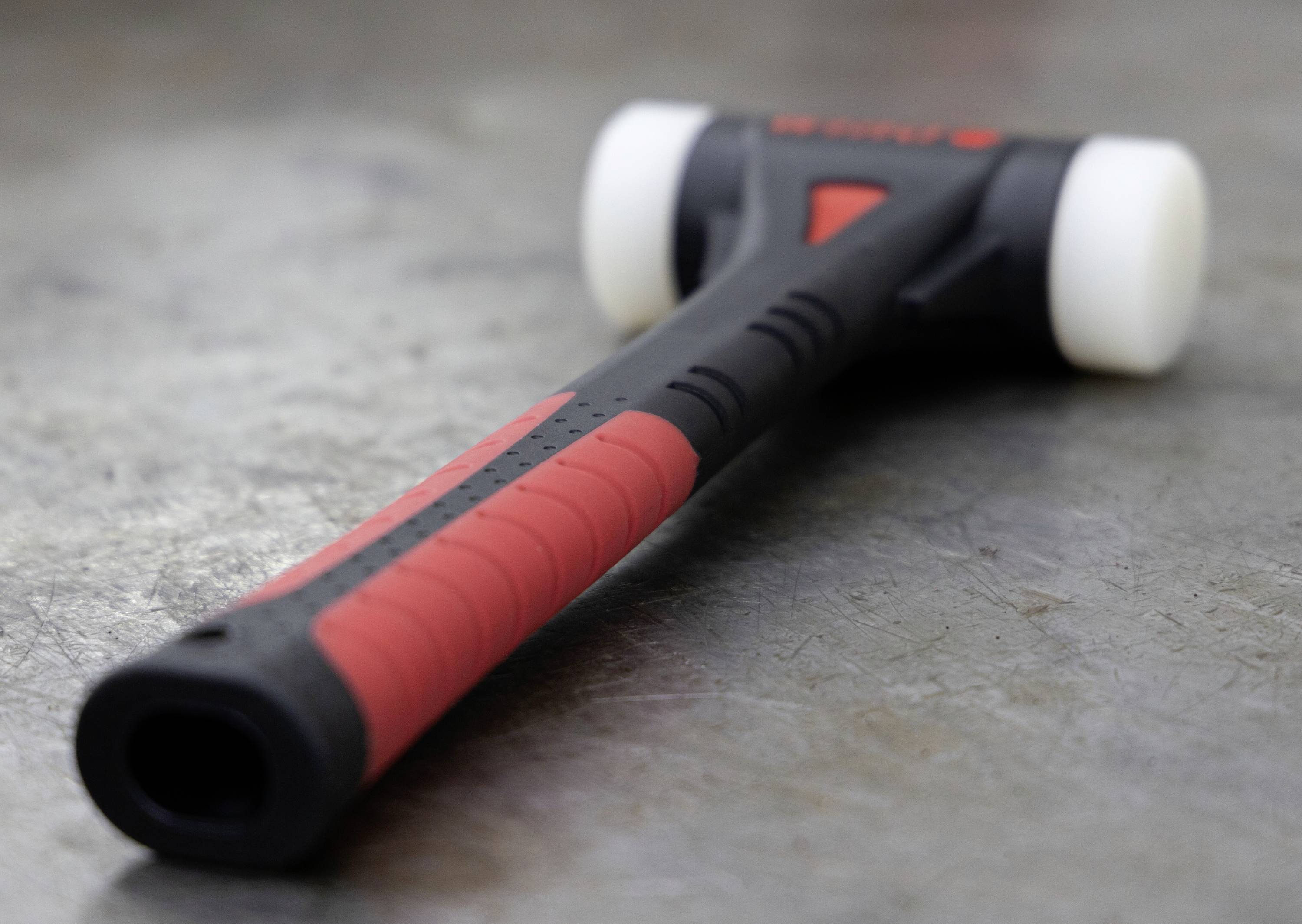 Młotek gumowy z czerwoną rączką i białymi powierzchniami uderzeniowymi leży na szarej metalowej powierzchni.