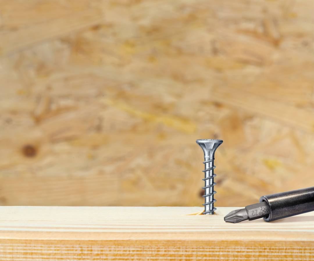Śrubokręt leży obok pionowo wbitej śruby na drewnianym podłożu z płyty sklejkowej.