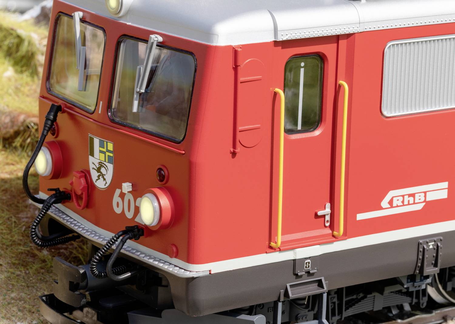 Czerwony pociąg z napisem 'RhB' i numerem '60' na torach, szczegółowy krajobraz makiety kolejowej w tle.