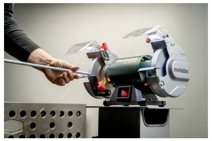 Osoba używa szlifierki stołowej Metabo do ostrzenia pręta, z latającymi iskrami. Maszyna jest zielona z osłonami ochronnymi na kołach.