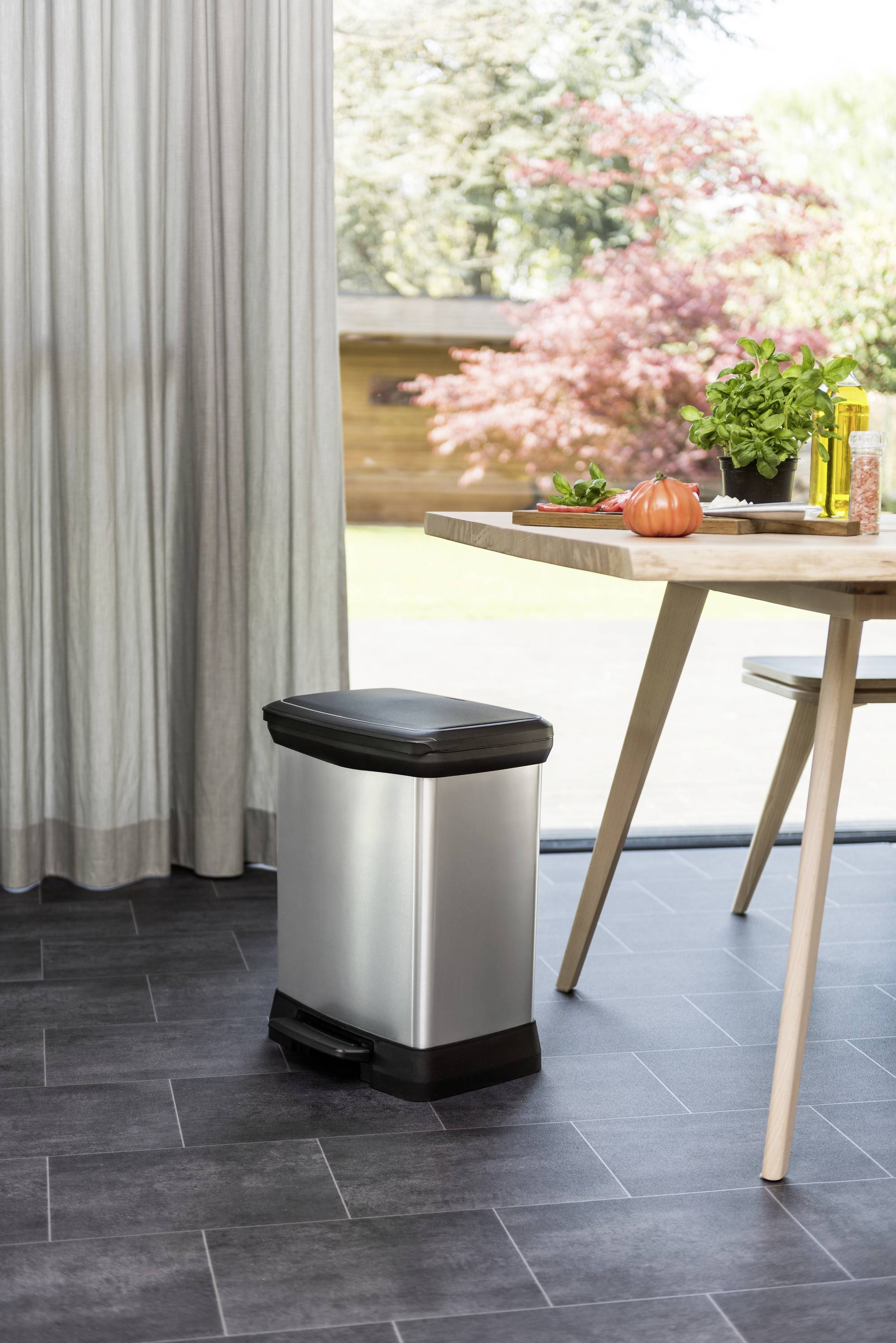 Srebrny kosz na śmieci z pedałem stoi obok stołu z roślinami i cebulą. W tle widać ogród.