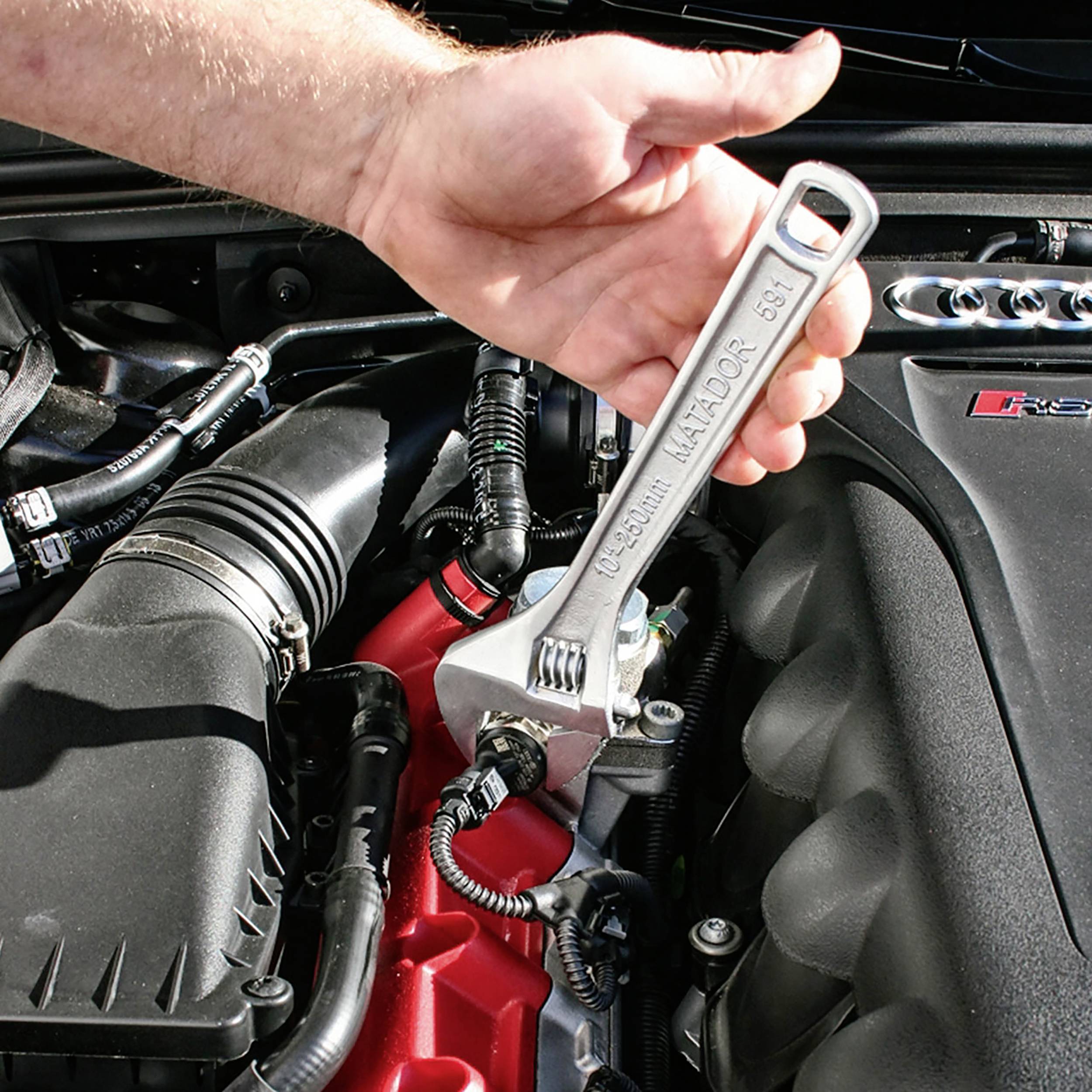 Jedna ręka trzyma klucz hydrauliczny i dokręca śrubę w komorze silnika samochodu. Silnik jest widoczny z wieloma kablami i elementami.