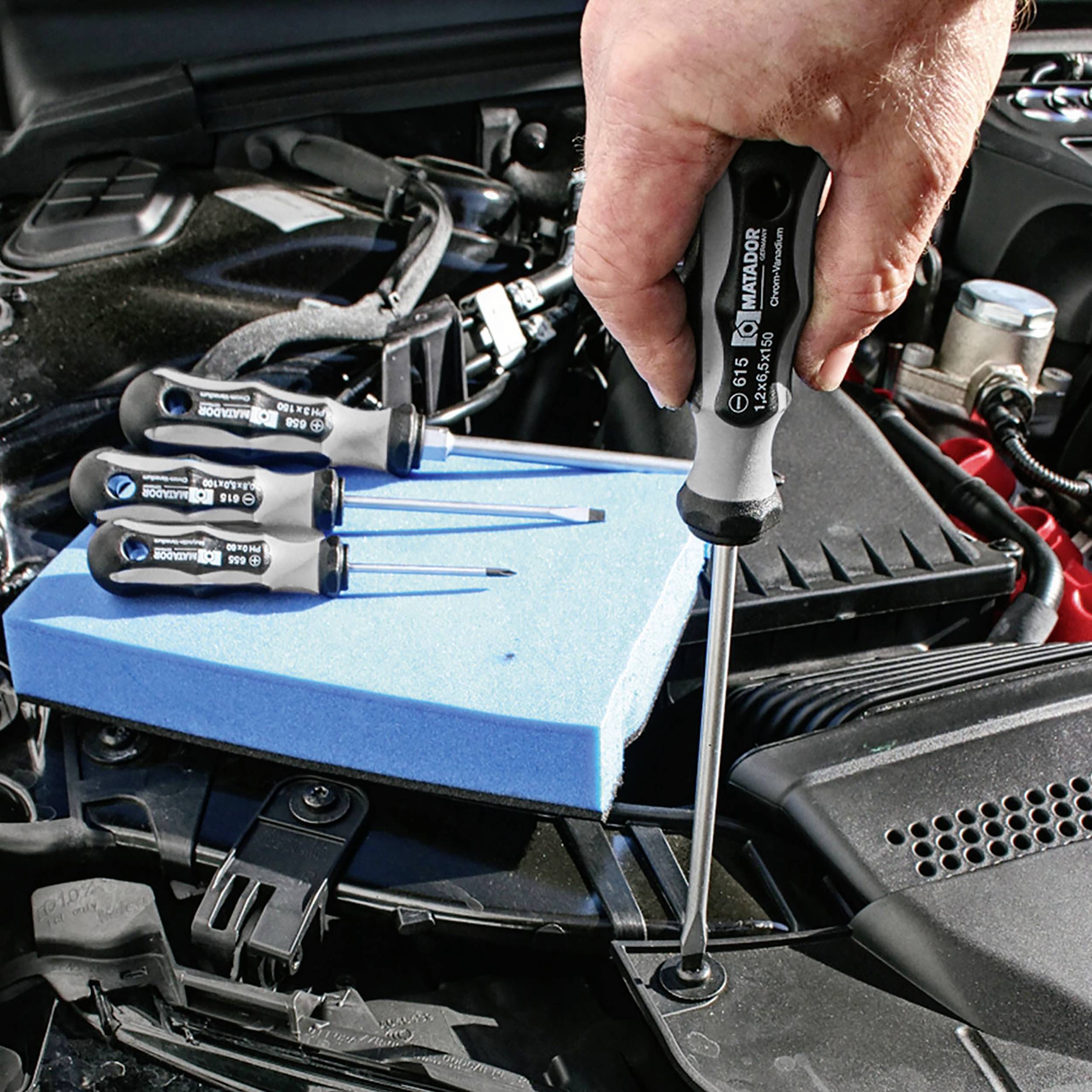 Jedna ręka trzyma śrubokręt nad otwartą obudową silnika. Trzy pozostałe śrubokręty leżą obok na niebieskiej gąbce.