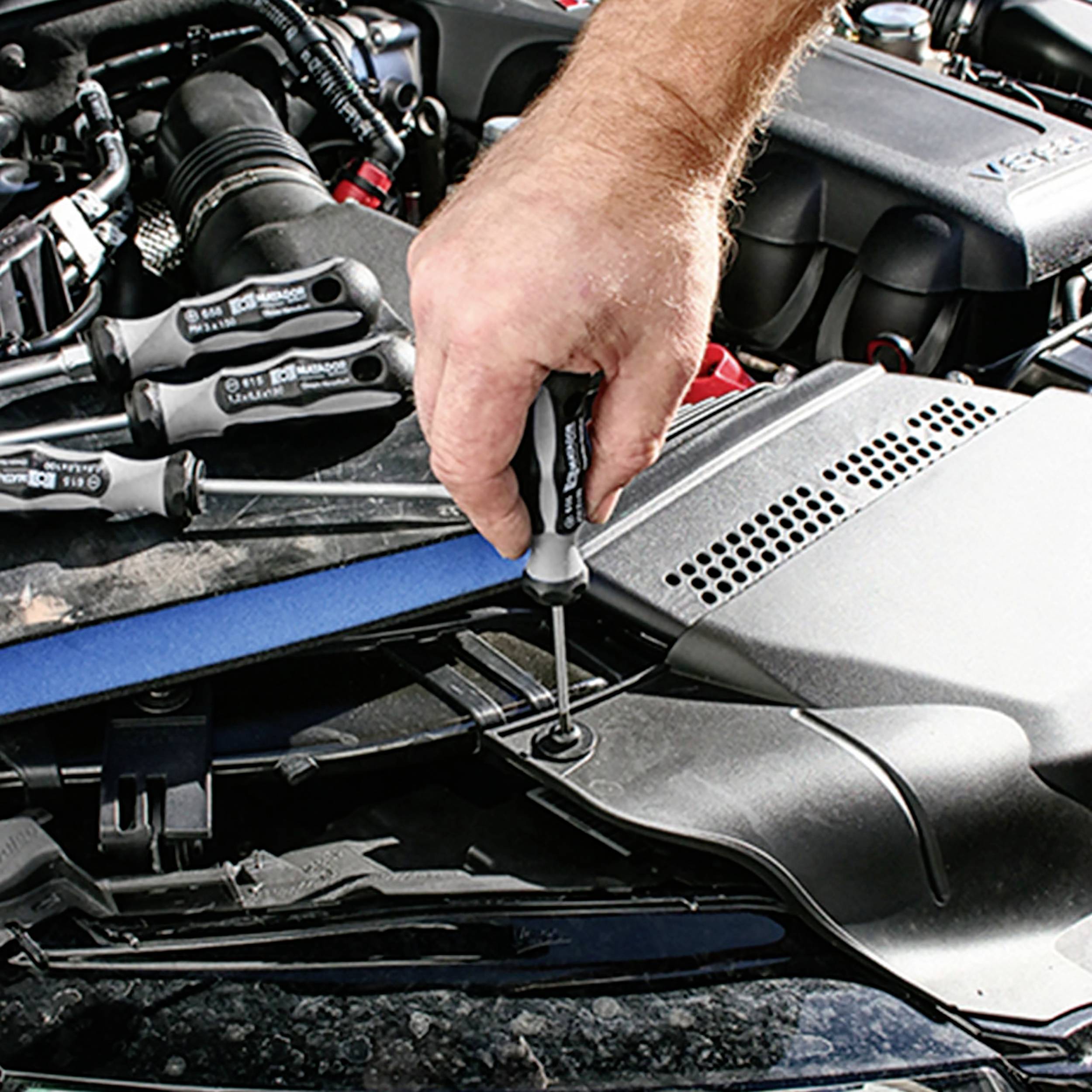 Osoba dokręca śrubę wkrętakiem w komorze silnika samochodu, wykonując czynności serwisowe.