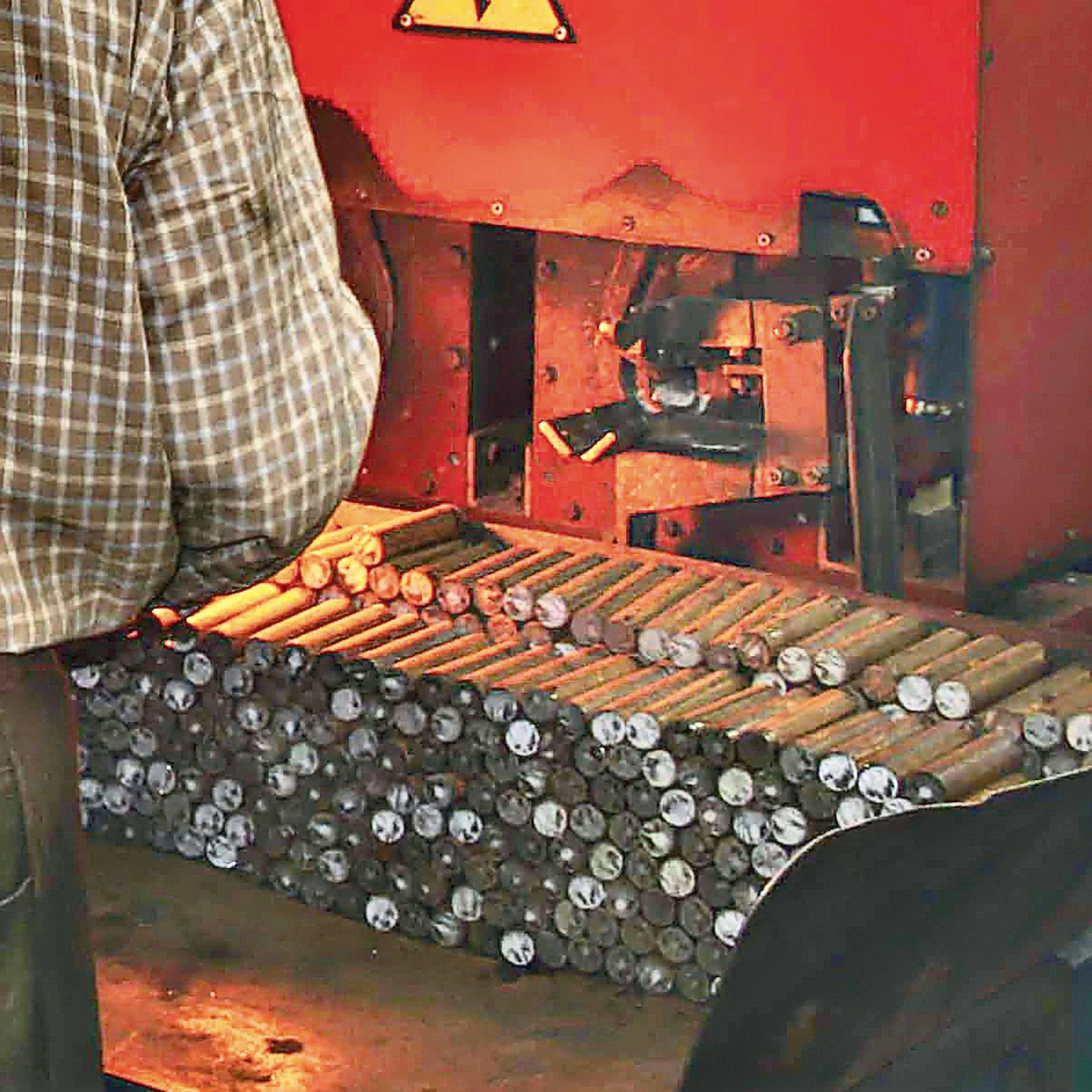 Mężczyzna w kratowanej koszuli stoi przed maszyną, która obrabia metalowe pręty. Pręty są ułożone w stos, gotowe do dalszej obróbki.