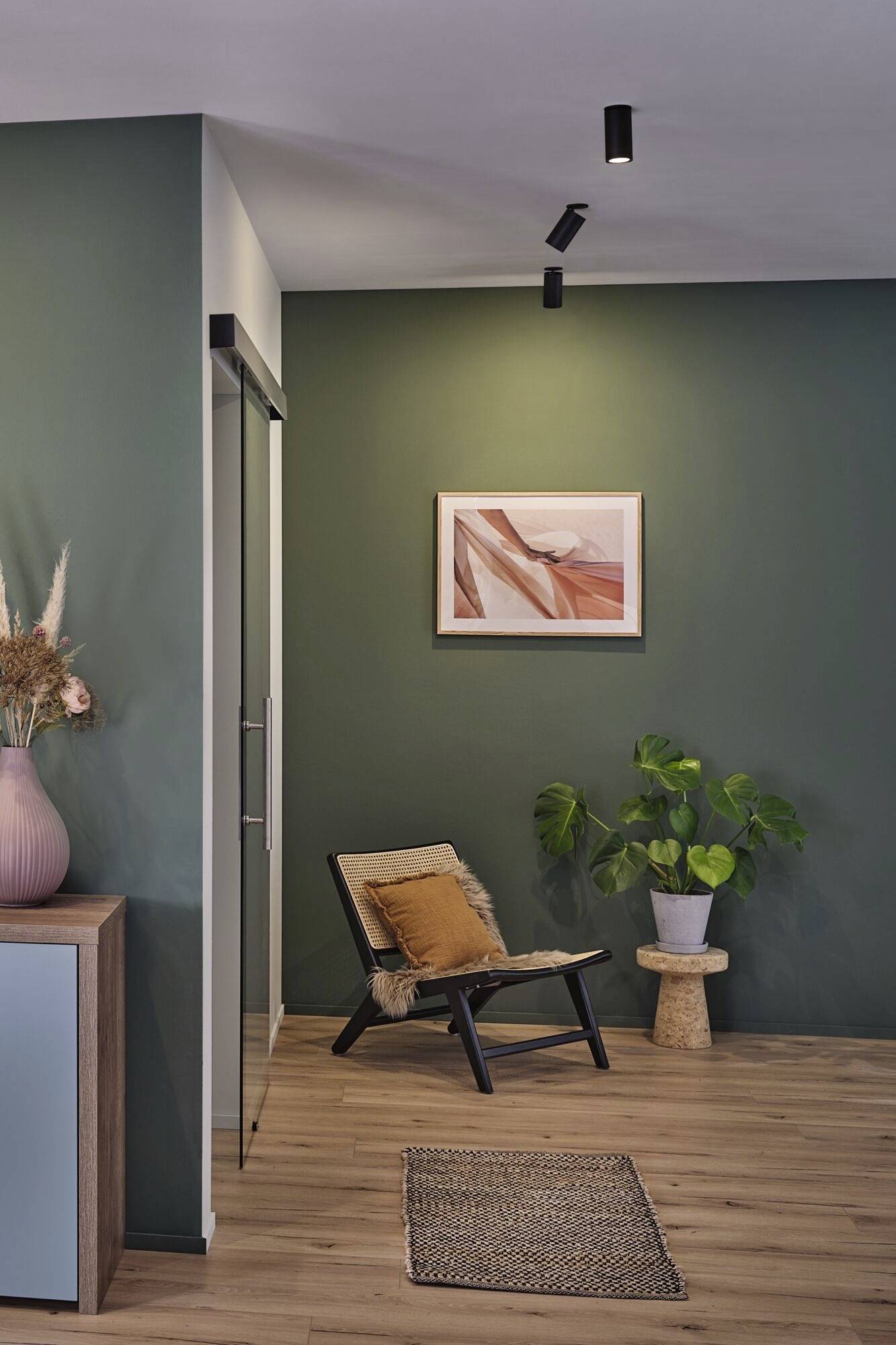 Nowoczesny salon z zielonymi ścianami, przytulnym fotelem, rośliną i oprawionym obrazem na ścianie.