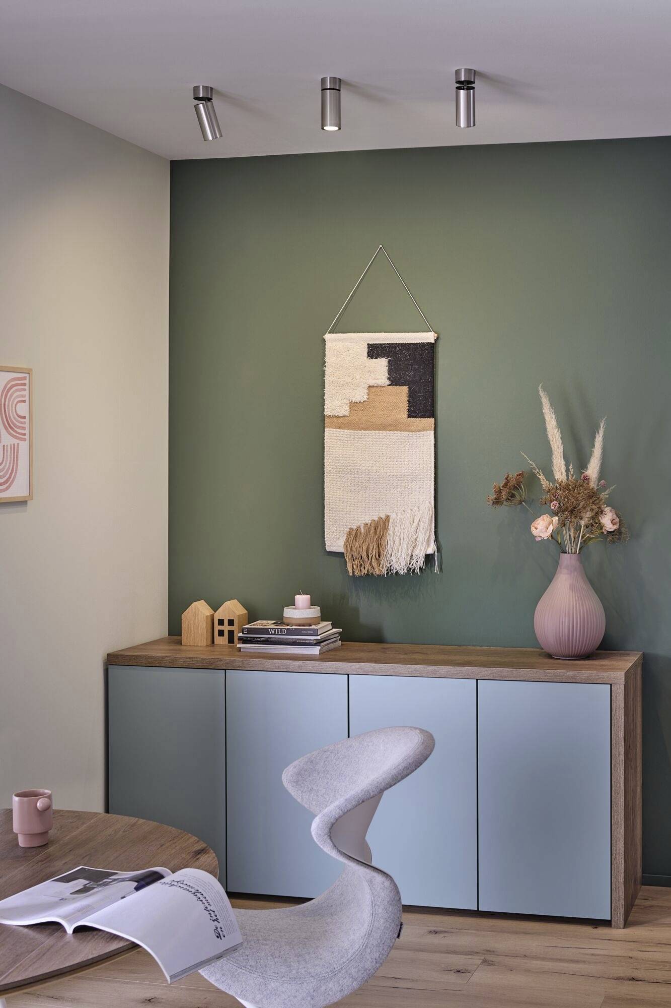 Nowoczesne wnętrze z zieloną ścianą, dekoracyjną tkaniną ścienną, drewnianymi meblami, wazonem z suszonymi kwiatami i designerskim krzesłem na pierwszym planie.