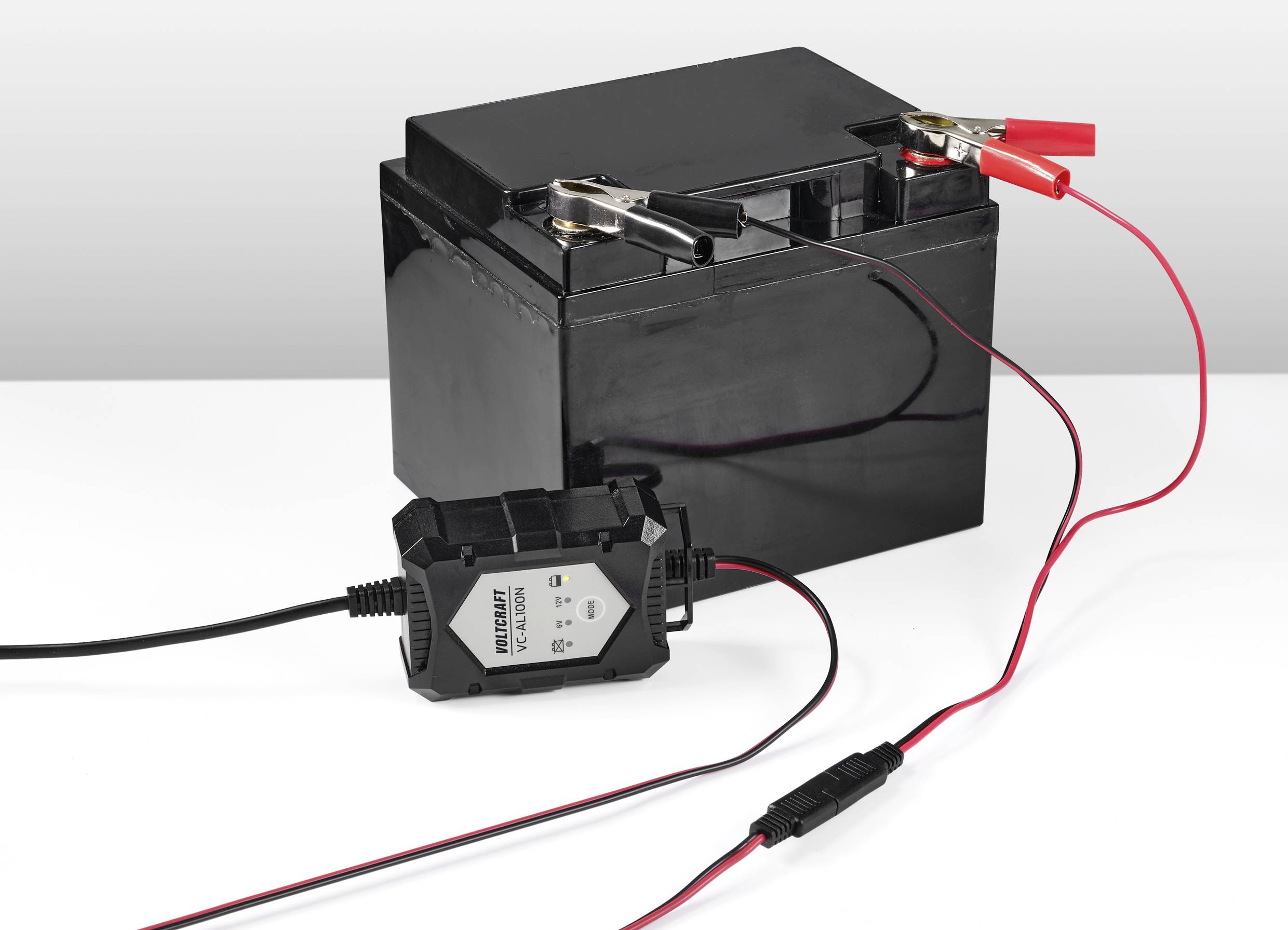 Czarna bateria samochodowa jest podłączona do ładowarki. Dwa przewody z czerwonymi i czarnymi zaciskami biegną od ładowarki do baterii.