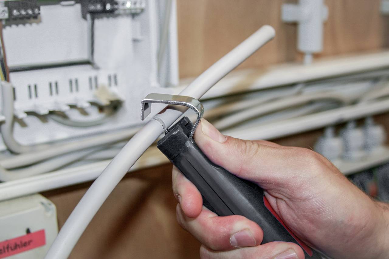 Osoba przycina białą wiązkę kabli za pomocą obcinacza w szafce rozdzielczej. W tle widoczne są elementy elektrotechniczne.