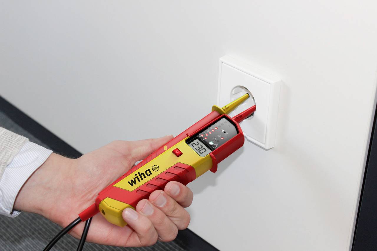 Osoba trzyma cyfrowy miernik napięcia przy gniazdku elektrycznym. Urządzenie wyświetla na ekranie wartość '230V'.