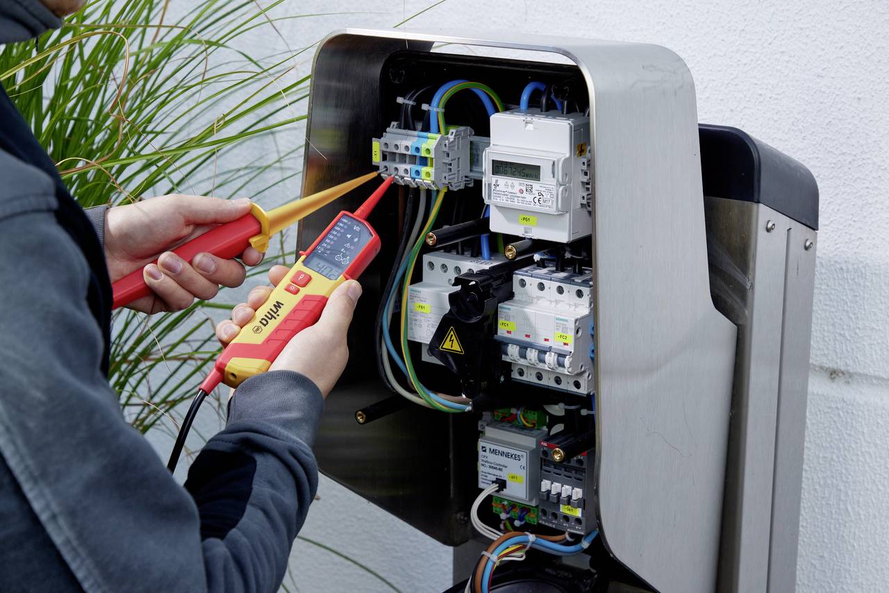 Osoba sprawdza elektryczne przyłącza w otwartej rozdzielni elektrycznej za pomocą multimetru. Widoczne są przewody i bezpieczniki.