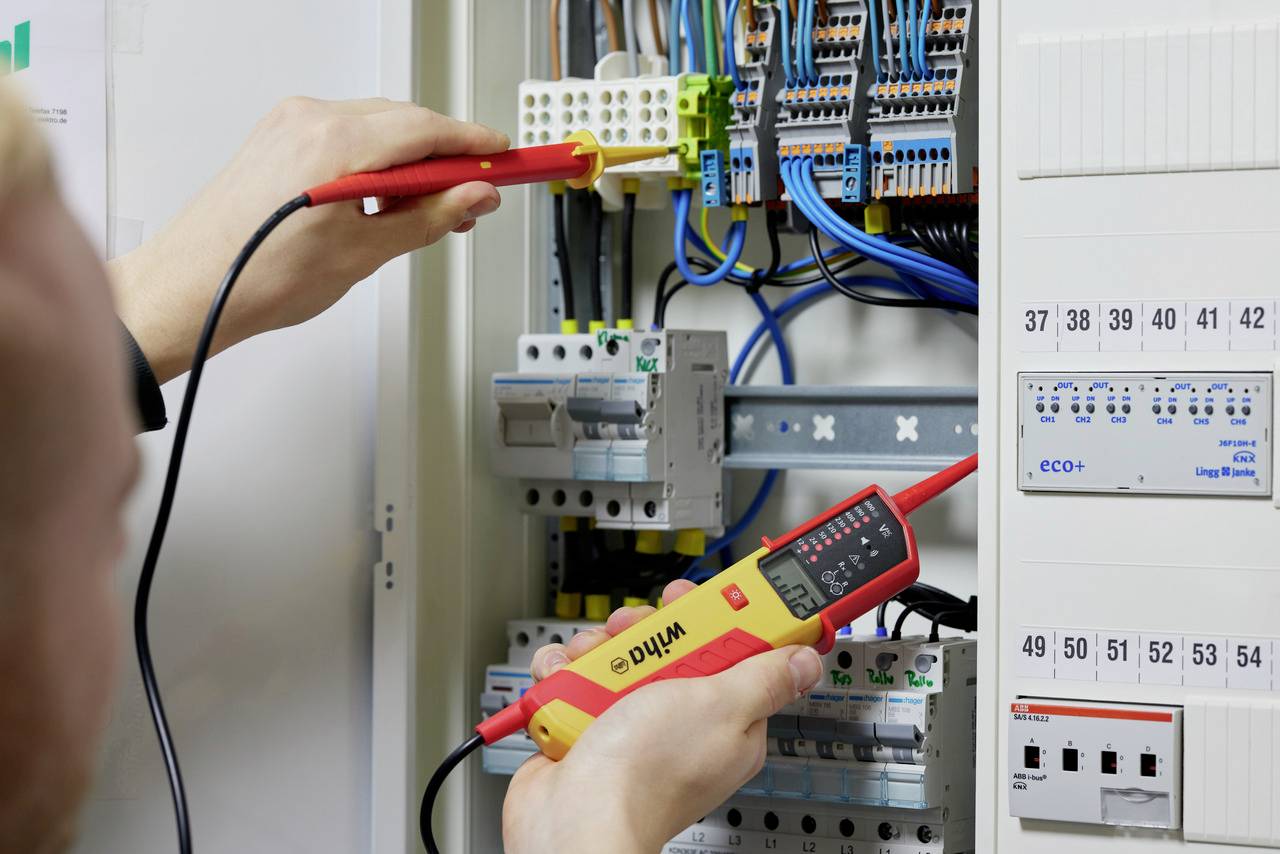 Osoba sprawdza kable w otwartej szafce sterowniczej za pomocą elektronicznego przyrządu pomiarowego. Widoczne są różnokolorowe przewody.