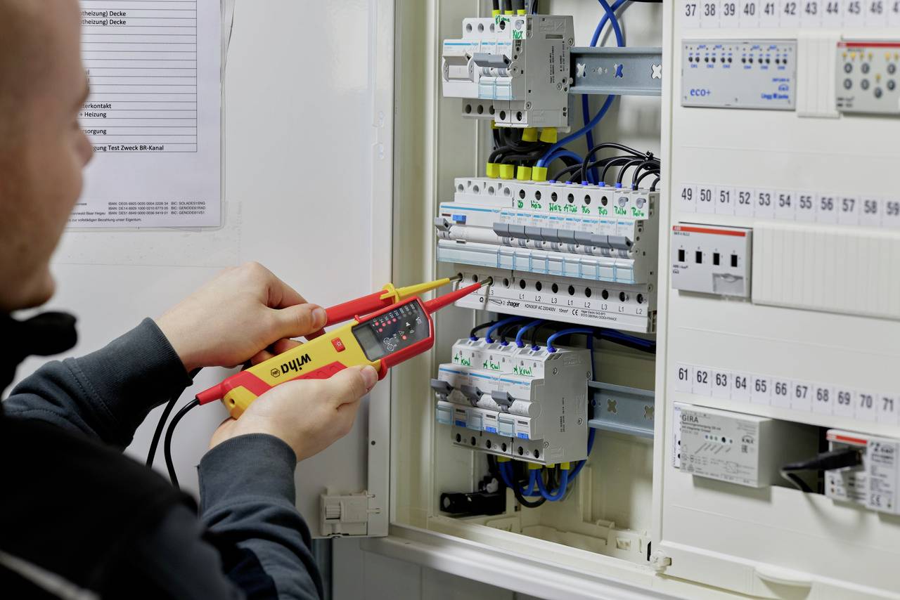 Osoba sprawdza instalację elektryczną w rozdzielnicy za pomocą próbnika napięcia. Główny nacisk kładziony jest na bezpieczeństwo i sprawność działania.