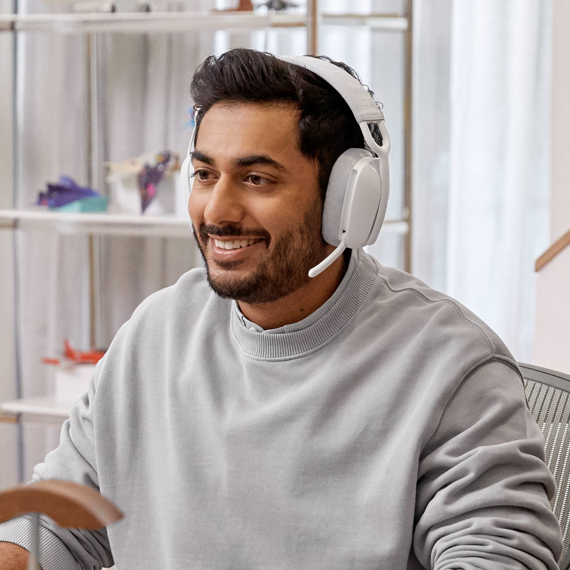 Mężczyzna w słuchawkach uśmiecha się i siedzi w jasnym pokoju. W tle widać półki z małymi dekoracjami.