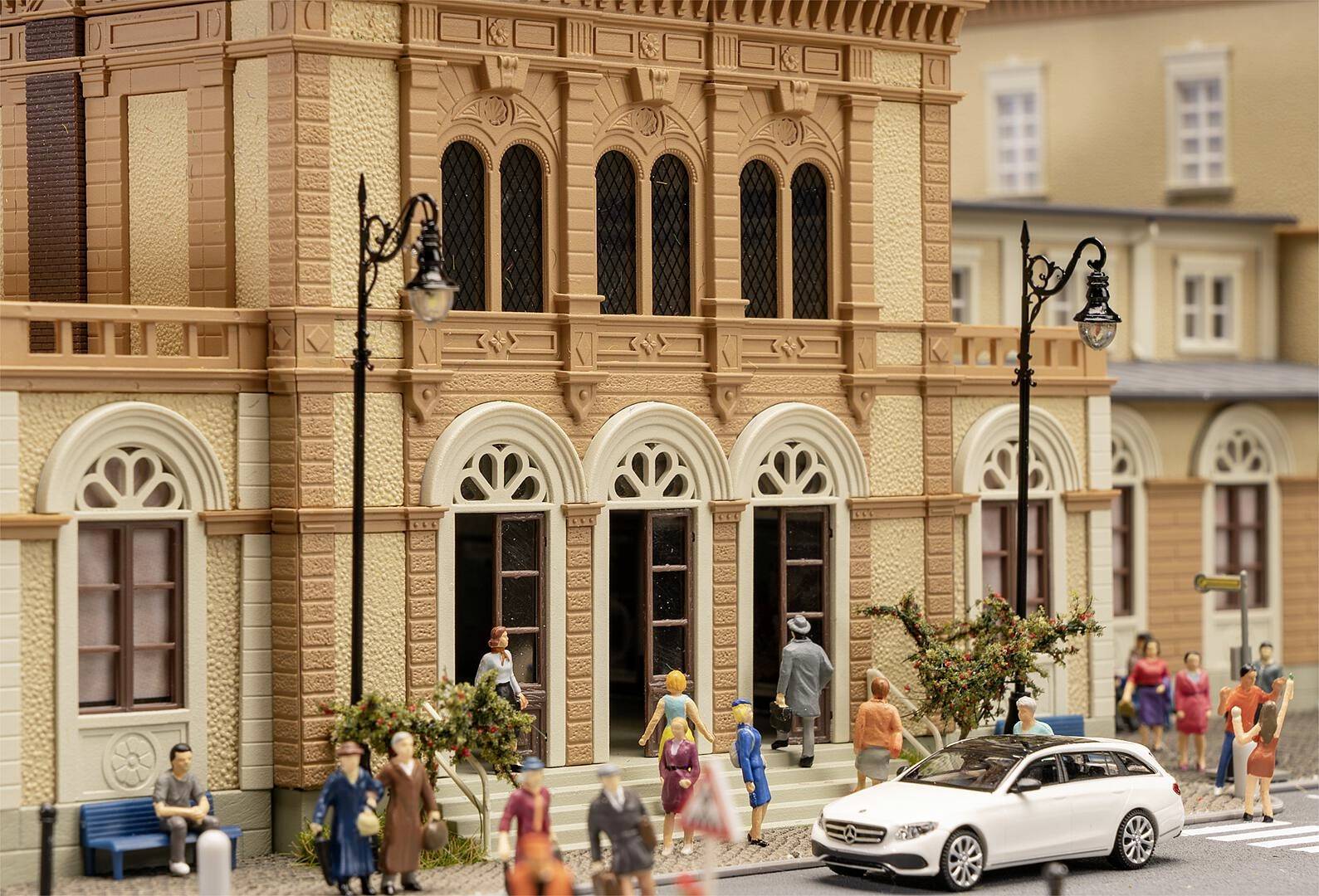 Miniaturowy model ożywionej sceny ulicznej przed zabytkowym budynkiem, z ludźmi i białym samochodem na pierwszym planie.