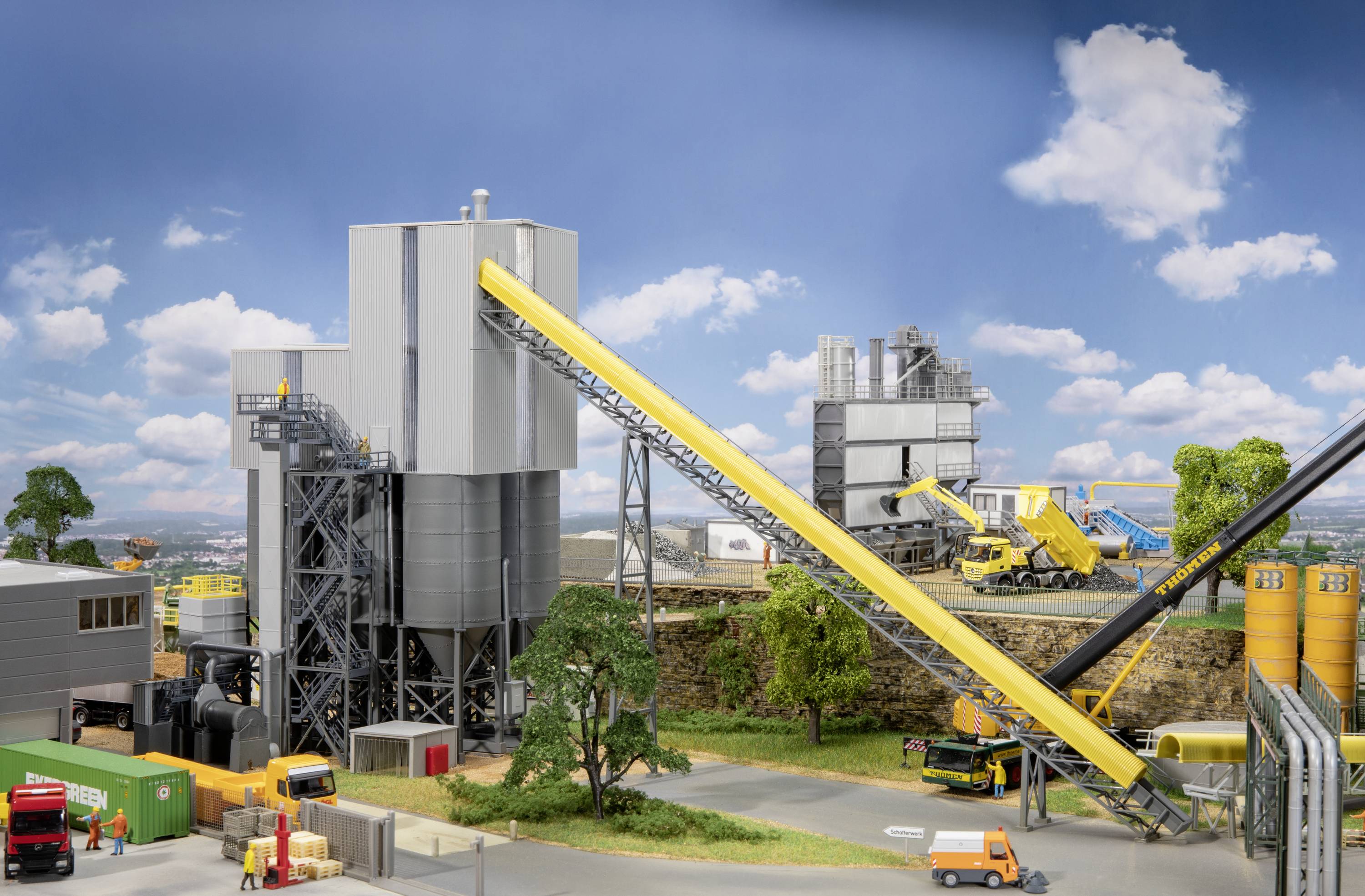 Przemysłowy zakład produkcyjny z silosami, przenośnikami taśmowymi i pojazdami budowlanymi. Na pierwszym planie zaparkowane są wielokolorowe pojazdy i kontenery.