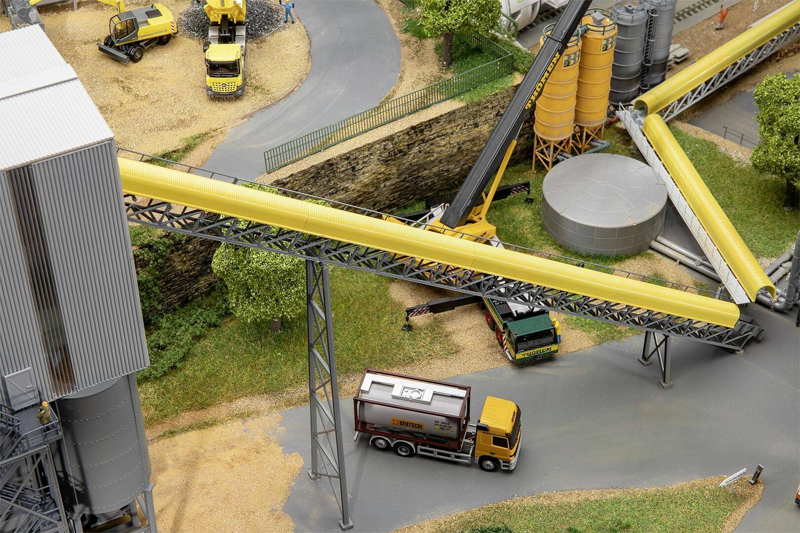 Miniaturowy model przedstawia scenę budowy z pojazdami, dźwigami i przenośnikami. Ciężarówka przejeżdża pod żółtym przenośnikiem taśmowym.