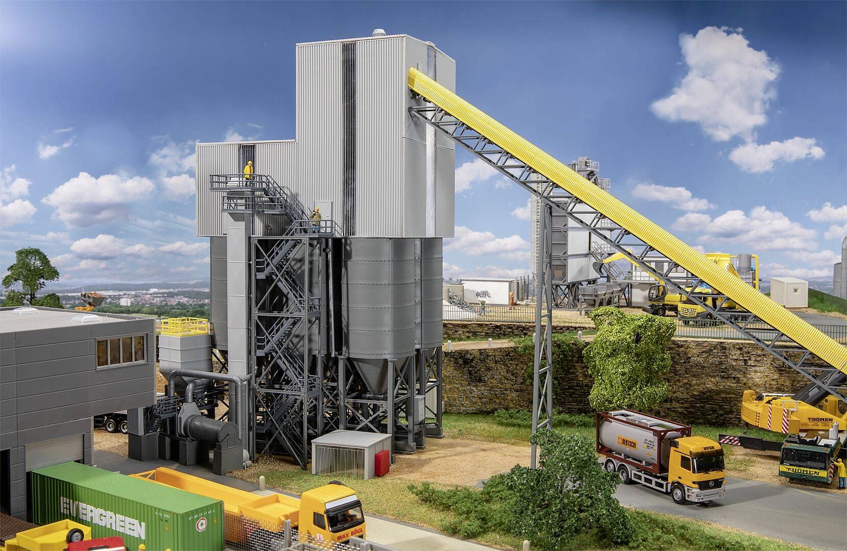 Stacja betoniarska z samochodami ciężarowymi na pierwszym planie pod czystym niebem. Obiekt posiada centralne budynki z przenośnikiem taśmowym.