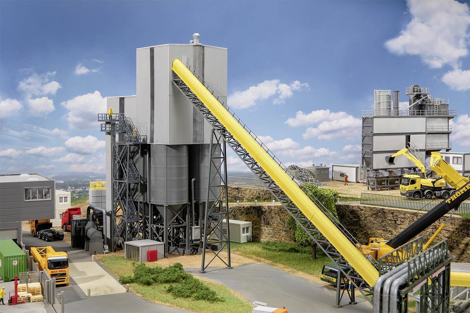 Teren przemysłowy z dużymi szarymi budynkami i żółtym przenośnikiem, otoczony ciężarówkami i dźwigiem, pod błękitnym niebem z nielicznymi chmurami.