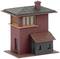 Mały, czerwono-brązowy model budynku z ciemnym dachem, kwadratowymi oknami i dobudówką po prawej stronie.