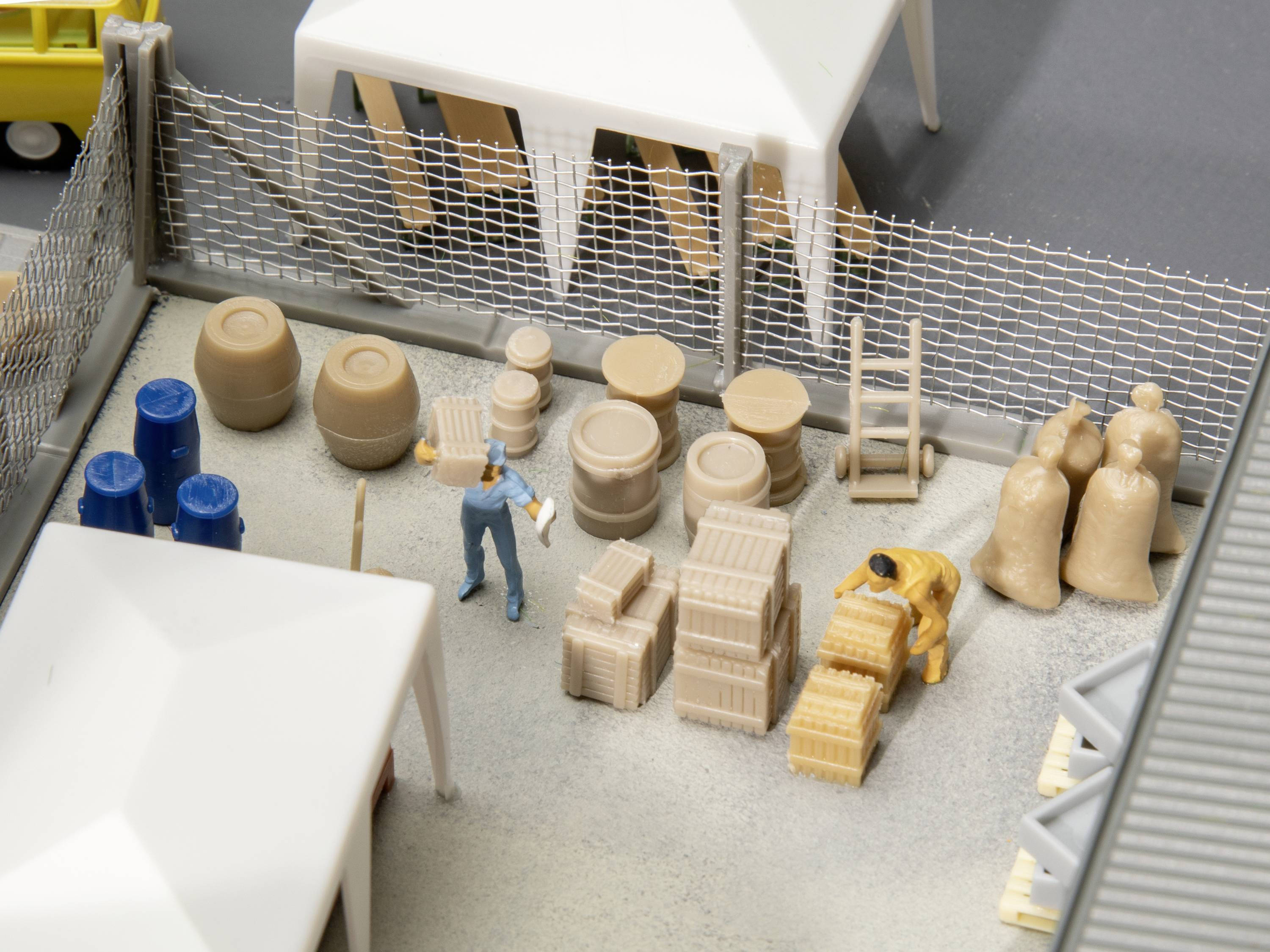 Miniaturowa scena magazynu z beczkami, workami i skrzyniami. Postać trzyma skrzynię przed białymi namiotami i ogrodzeniem drucianym.