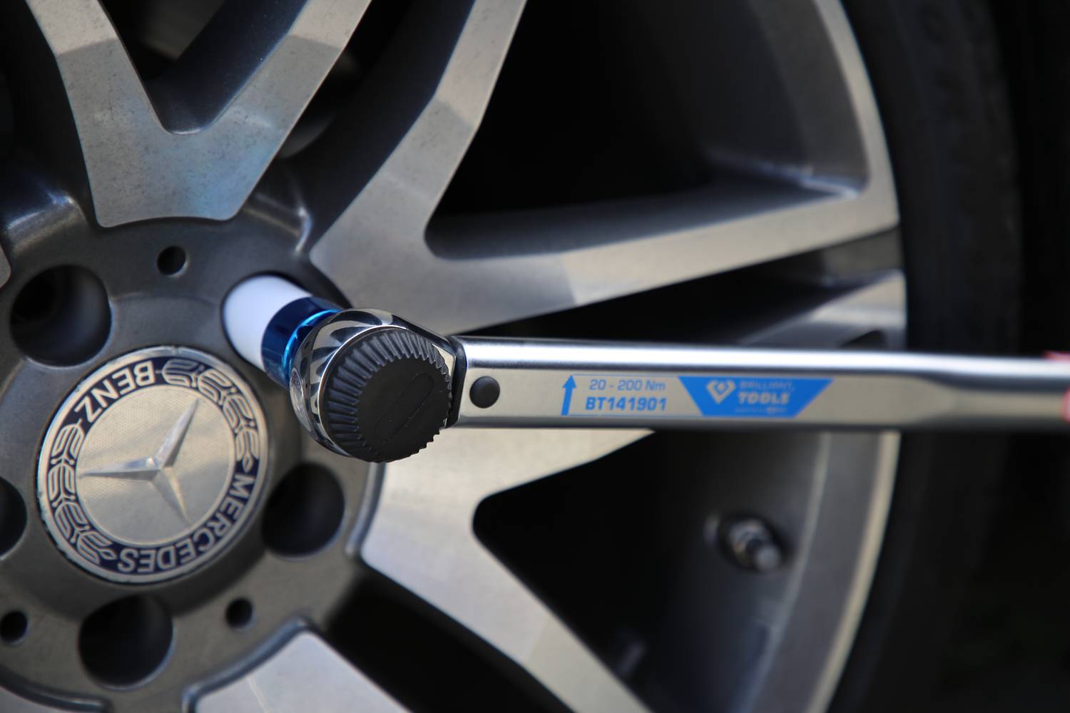 Klucz dynamometryczny dokręca nakrętki koła samochodowego. Koło ma srebrne szprychy i kołpak z logo firmy.