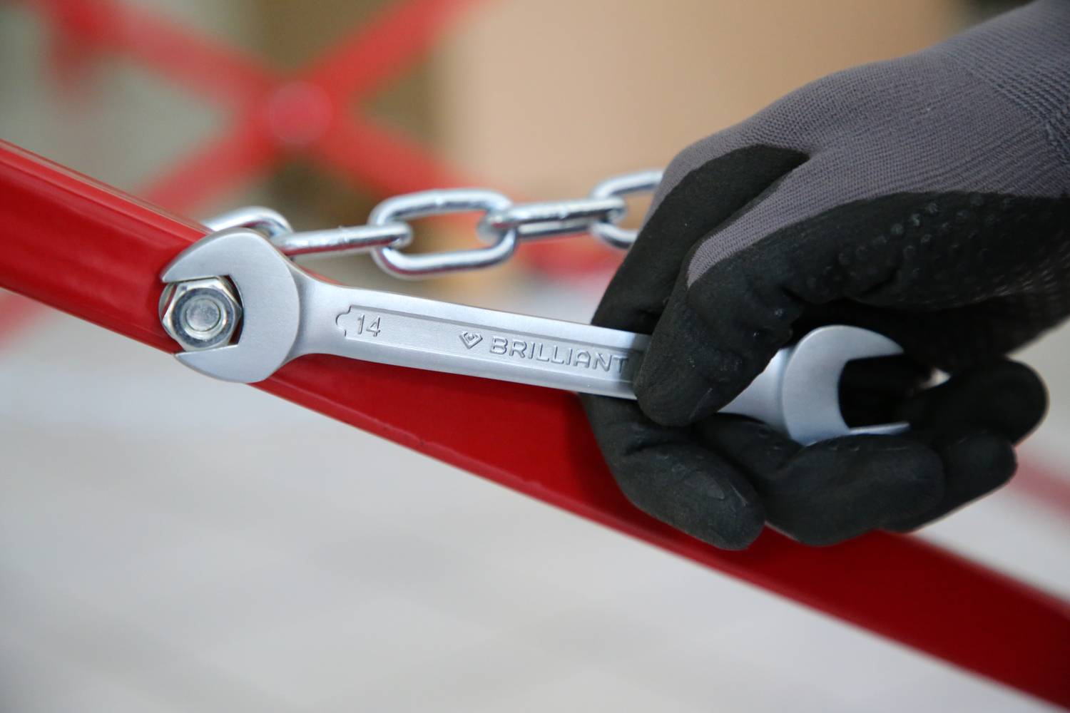 Rękawica trzyma klucz nastawny, który dokręca śrubę na czerwonej metalowej ramie. Łańcuch jest przymocowany do ramy.