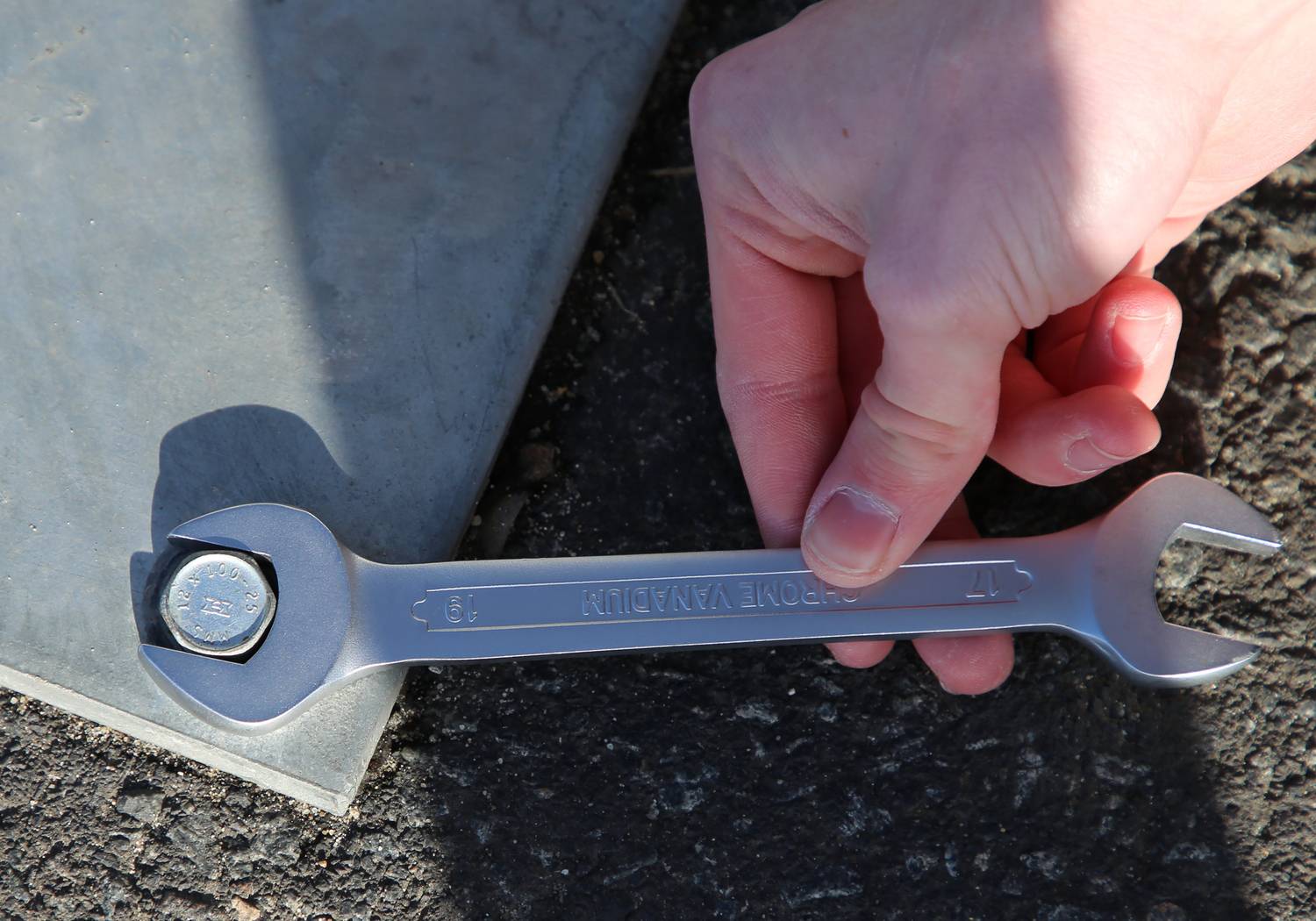 Jedna ręka trzyma klucz płaski, który przytrzymuje śrubę, na asfaltowej powierzchni obok metalowej płyty.