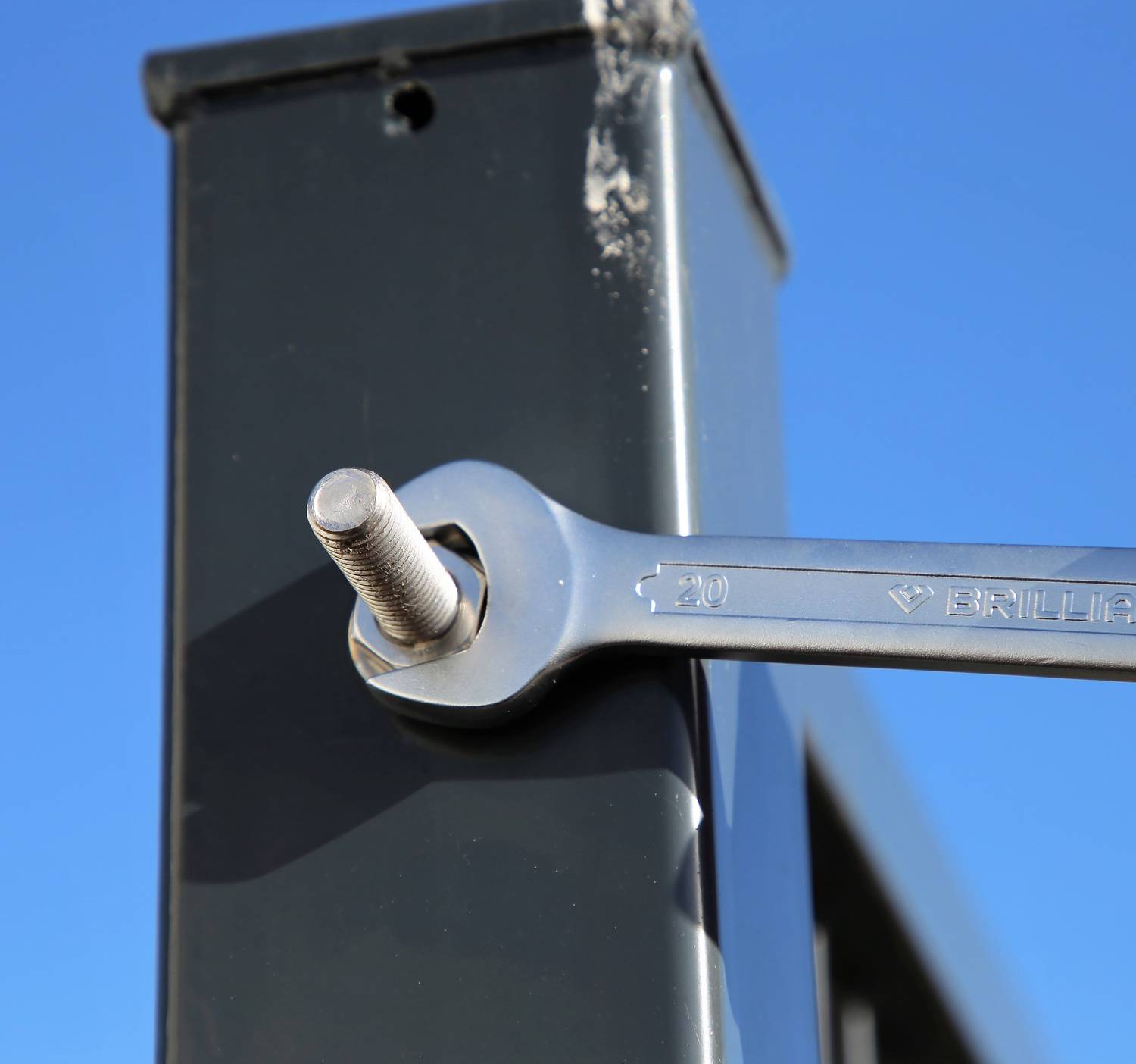 Klucz płaski dokręca śrubę na metalowej ramie. Na tle niebo jest błękitne, bez żadnych innych widocznych szczegółów.