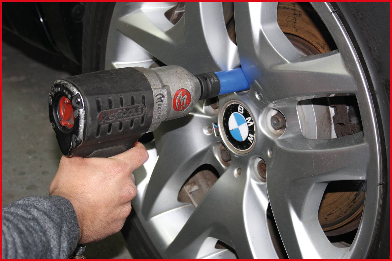 Ręka obsługuje pneumatyczny klucz udarowy, dokręcając śrubę w oponie samochodu z logo BMW.