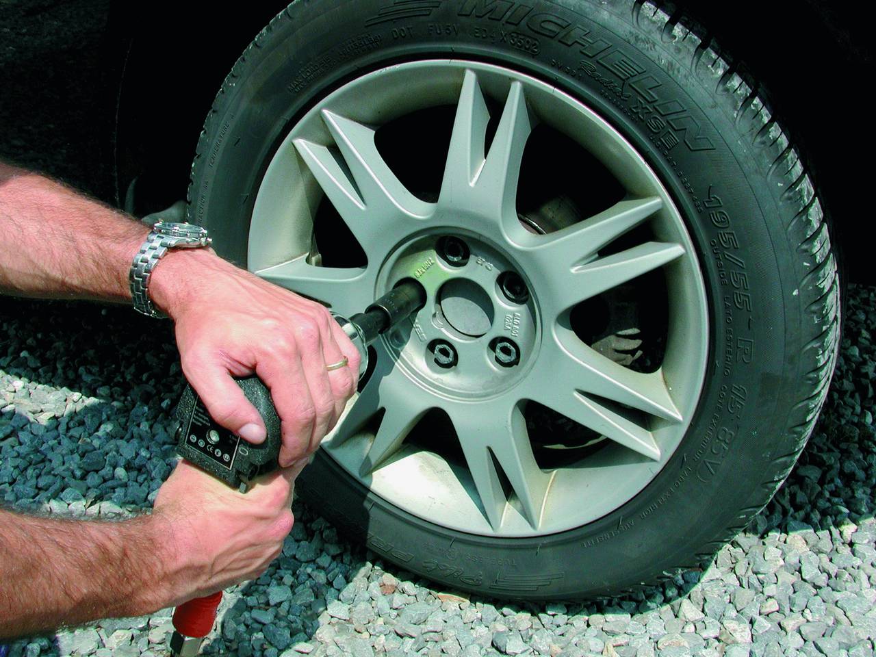 Osoba dokręca śruby kół samochodu wkrętarką udarową na żwirowej powierzchni.