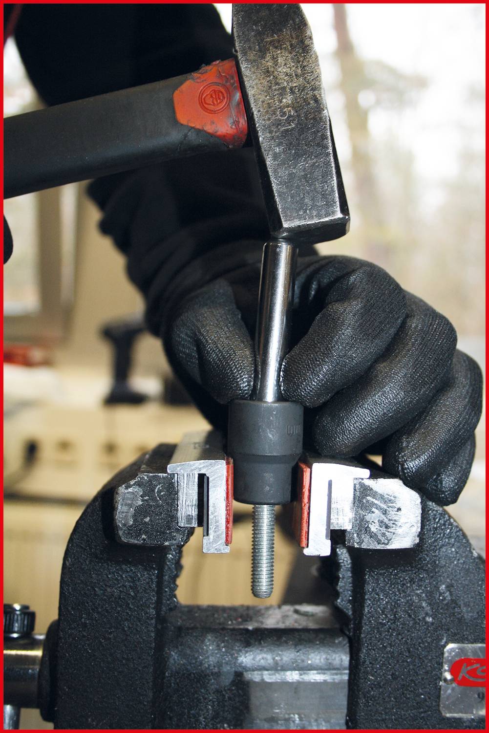 Dłoń trzyma metalowy pręt w imadle, podczas gdy młotek go obrabia. W tle widoczne jest środowisko warsztatu.