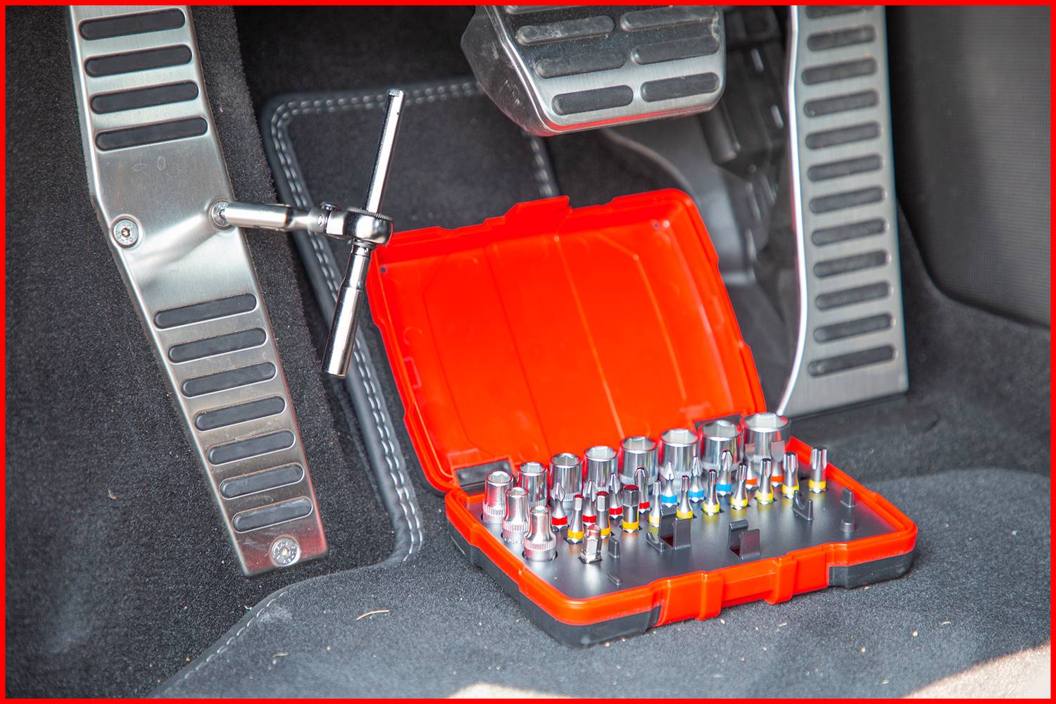 Otwarty czerwony zestaw narzędziowy z nakładkami leży nad obszarem pedałów samochodu.