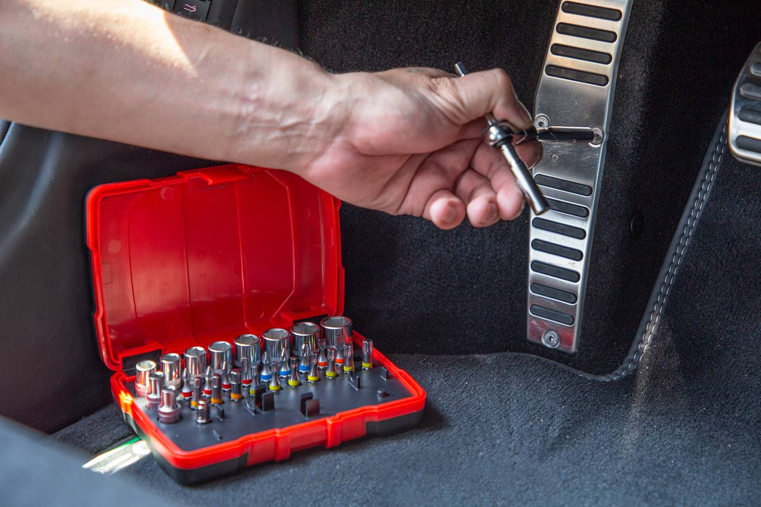 Ręka używa narzędzia do wkręcania śruby w samochodzie. Obok na podłodze leży czerwony zestaw narzędziowy.