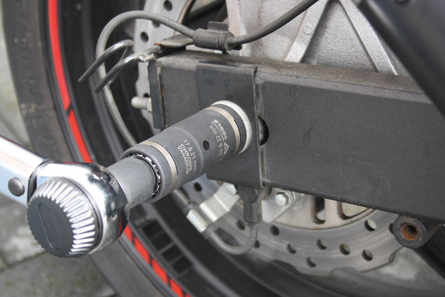 Zbliżenie motocykla z kluczem nasadzonym na nakrętkę osi. W tle widoczna tarcza hamulcowa i bieżnik opony.