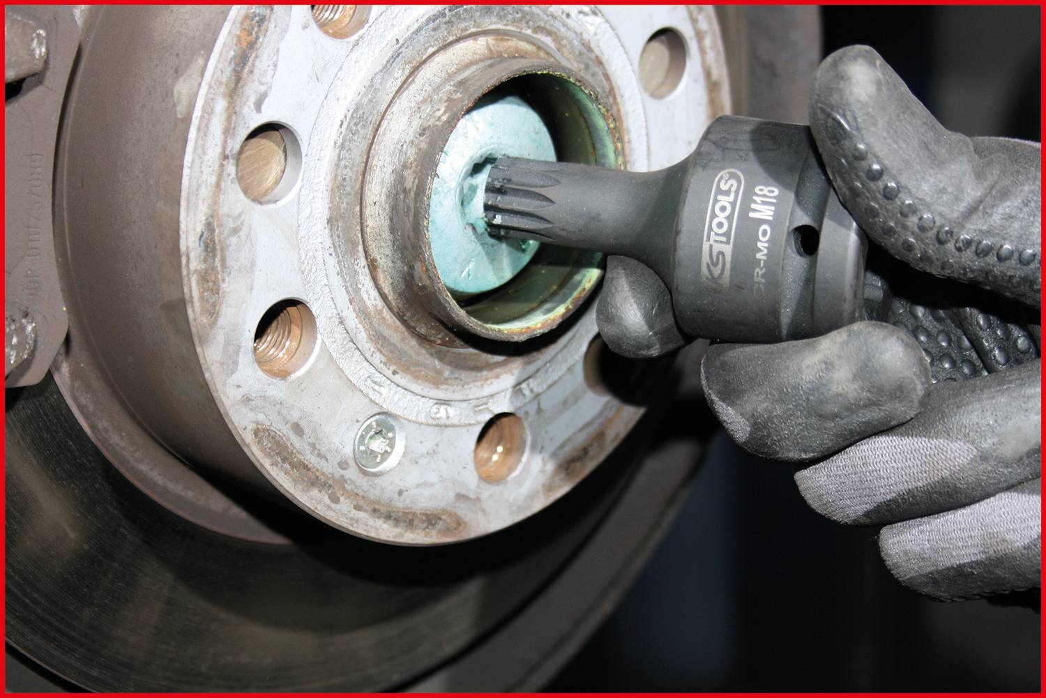 Dłoń w roboczej rękawicy trzyma końcówkę narzędziową i pracuje przy części samochodowej, prawdopodobnie przy tarczy hamulcowej.