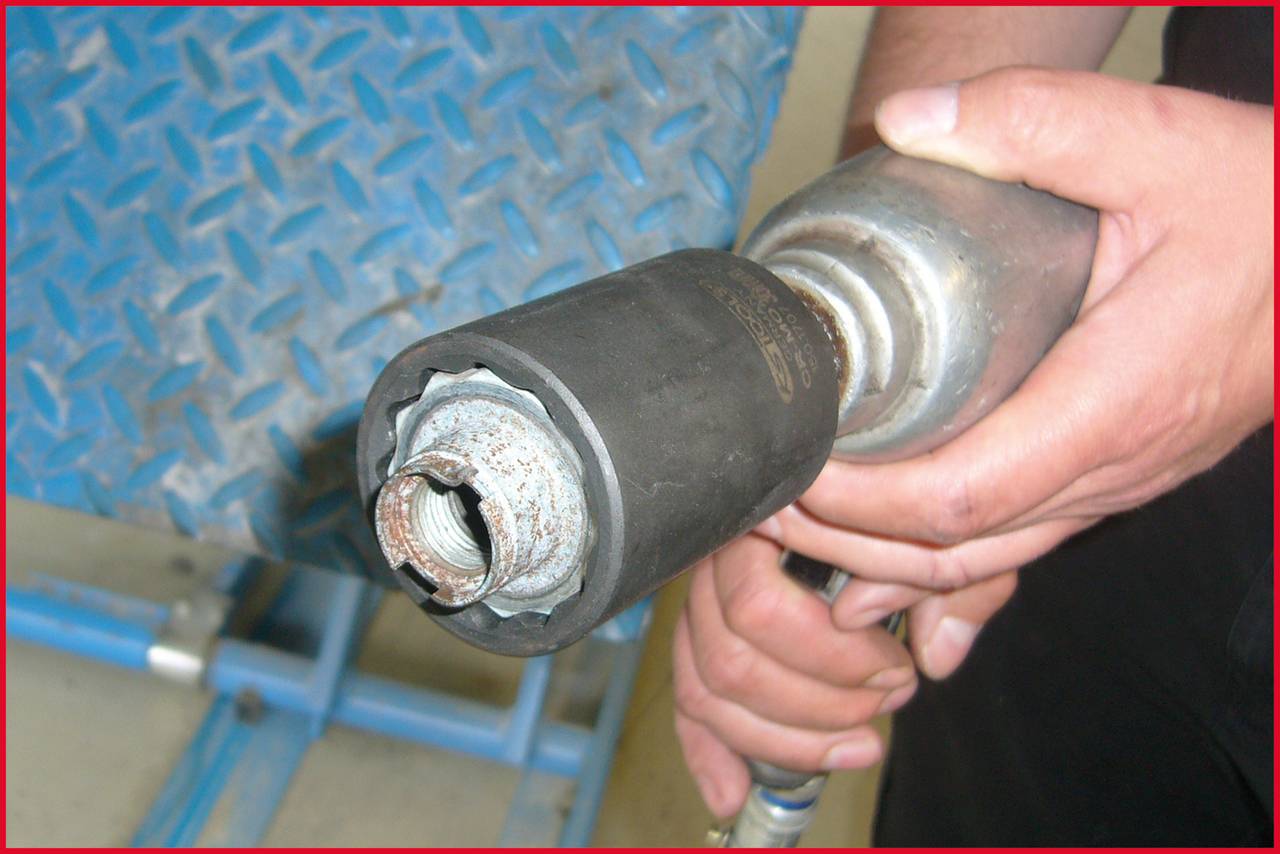 Osoba trzyma mechaniczny klucz udarowy. Na tylnym planie widoczna jest niebieska, metaliczna powierzchnia o wzorze diamentowym.