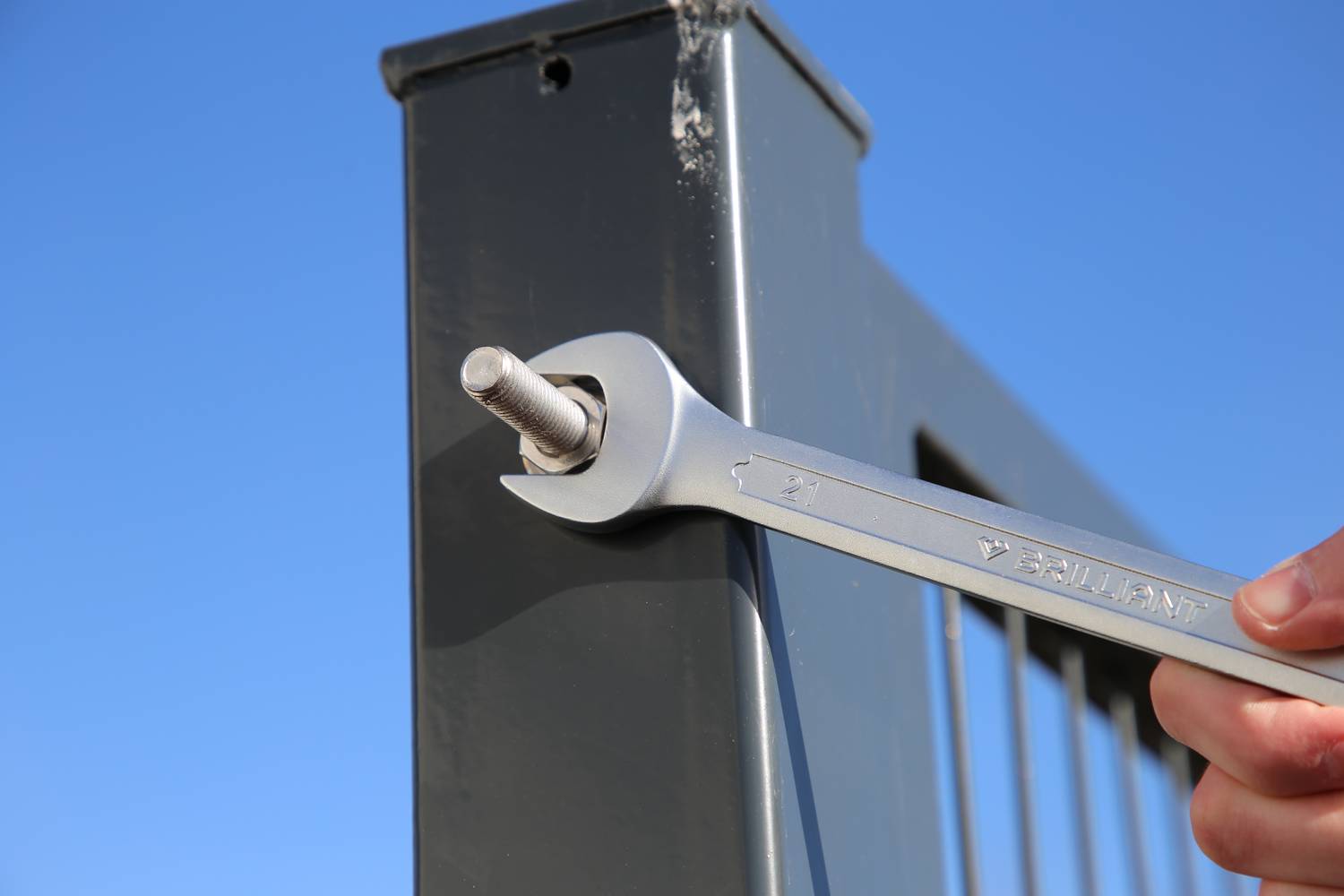Dłoń dokręca śrubę kluczem na metalowym słupie, podczas gdy w tle niebo jest błękitne.