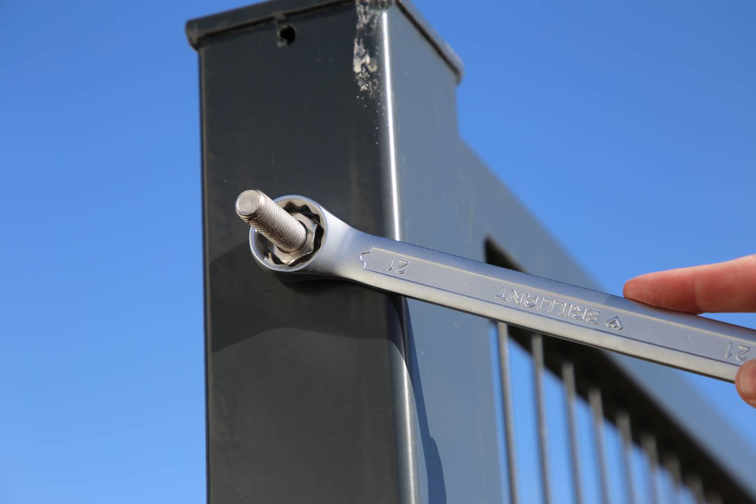 Klucz nastawny dokręca śrubę na szarym metalowym poręczu. W tle czyste, błękitne niebo.