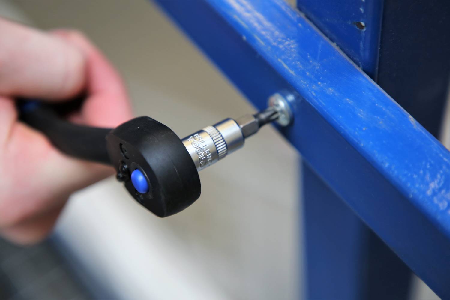 Ręka wkręca wkręt niebieskim śrubokrętem w niebieską strukturę metalową.