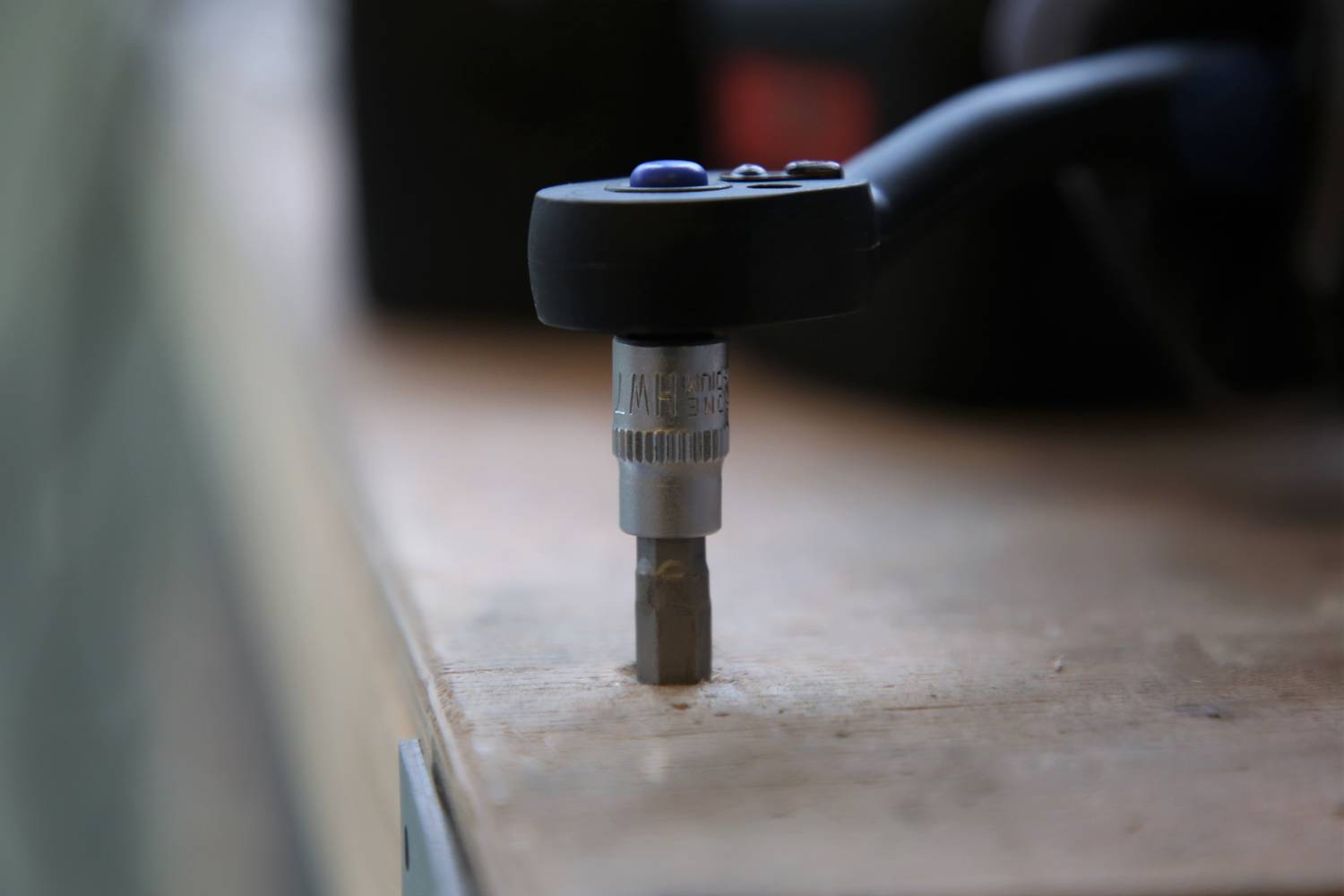 Wkrętarka akumulatorowa z osadzonym bitem leży na stole warsztatowym. Ostrość ujęcia skierowana na bit i szybkozaciskowy uchwyt. Otoczenie narzędziowe.