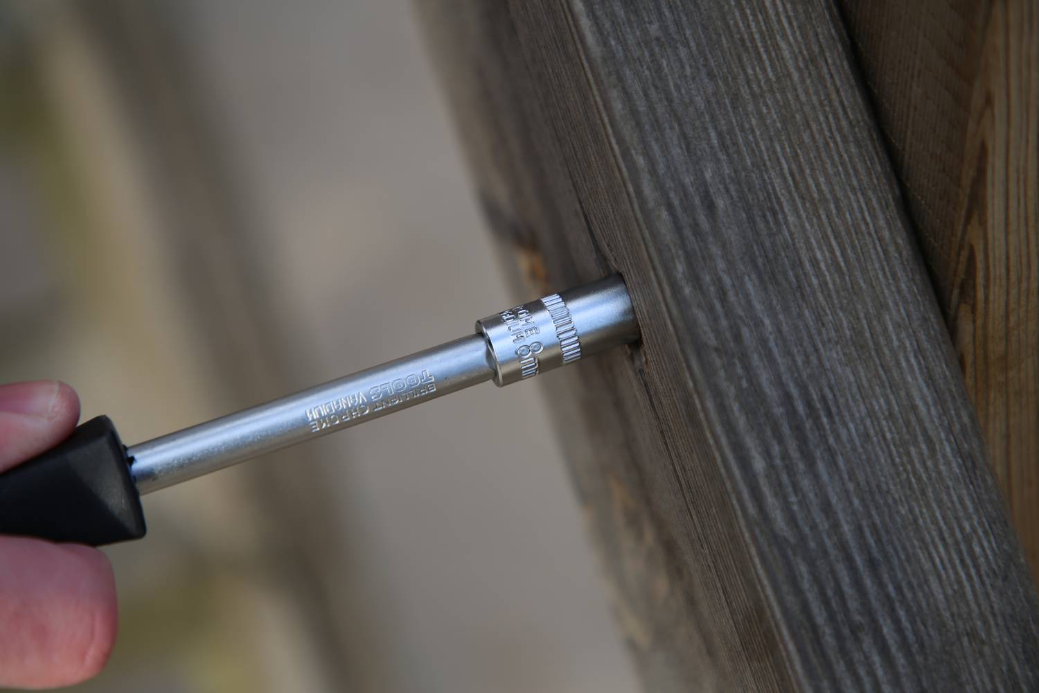 Osoba mierzy głębokość otworu wiertniczego w drewnianym belku za pomocą głębokościomierza. Belka wygląda na ciemnobrązową i ma wyraźną teksturę.